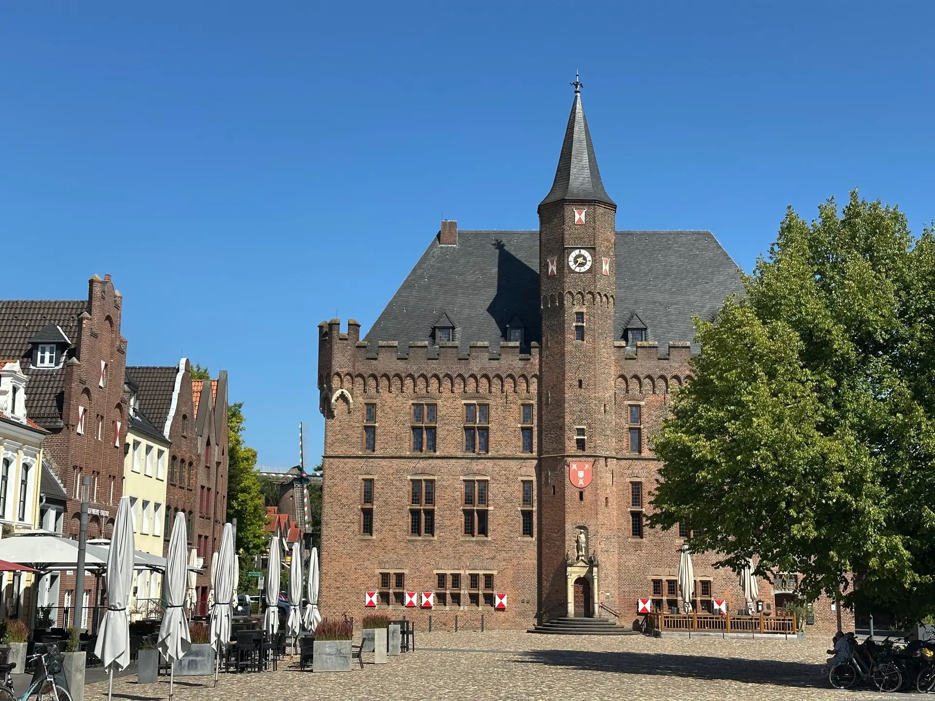 Backsteinbau mit Schieferdach, Zinnen und Turm, davor ein Baum, im Hintergrund eine Mühle