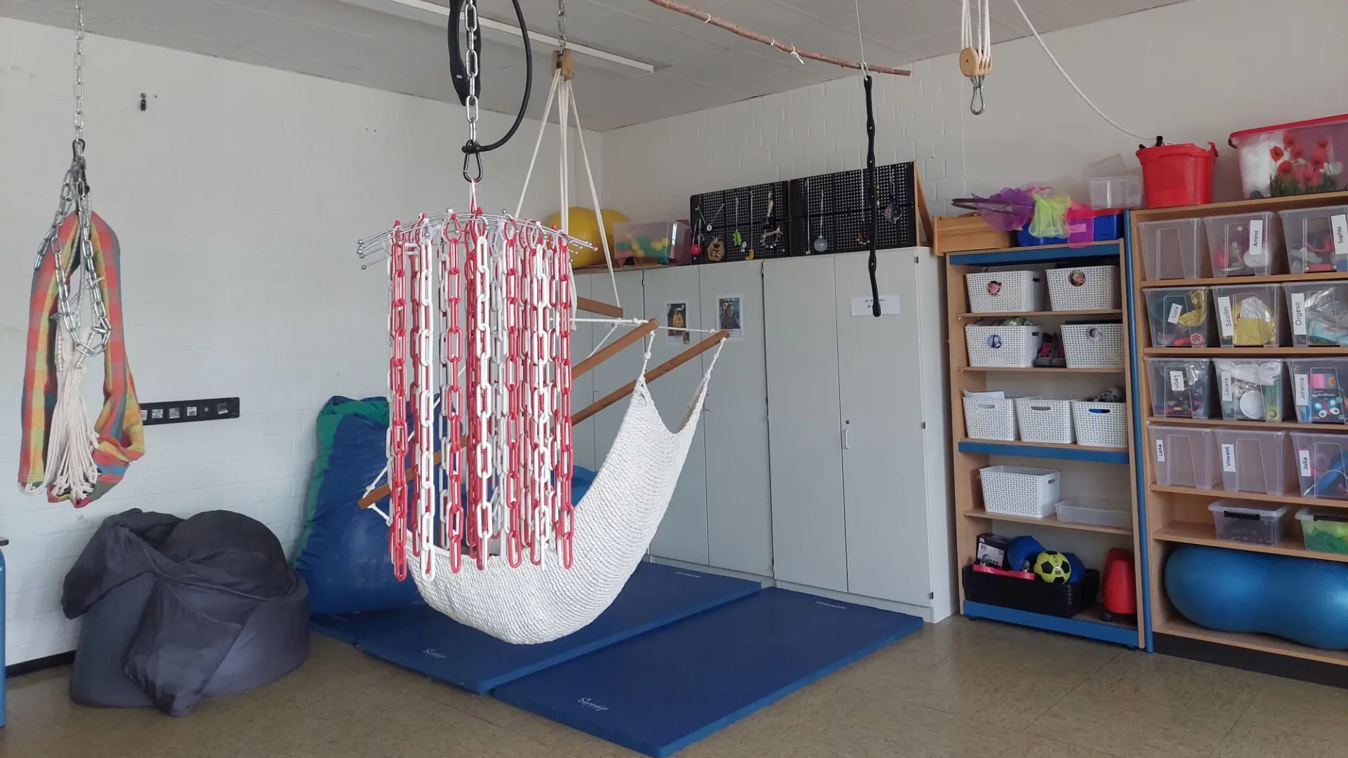 Ein Raum mit einer Hängematten-Schaukel, Kissen und Gymnastikmatten – an der Wand Schränke und Regale mit bunten Materialen zum Fühlen und Entdecken