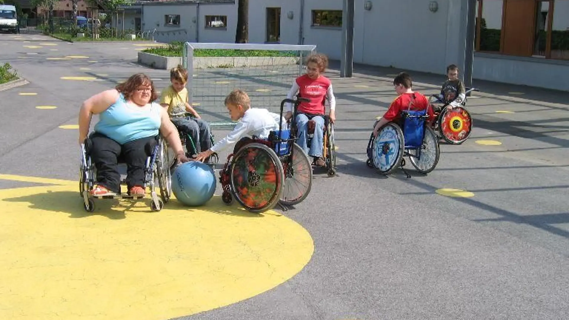 Auf einem mit gelben Kreisen bemalten Schulhos spielen mehre Kinder im Rollstuhl mit einem Ball