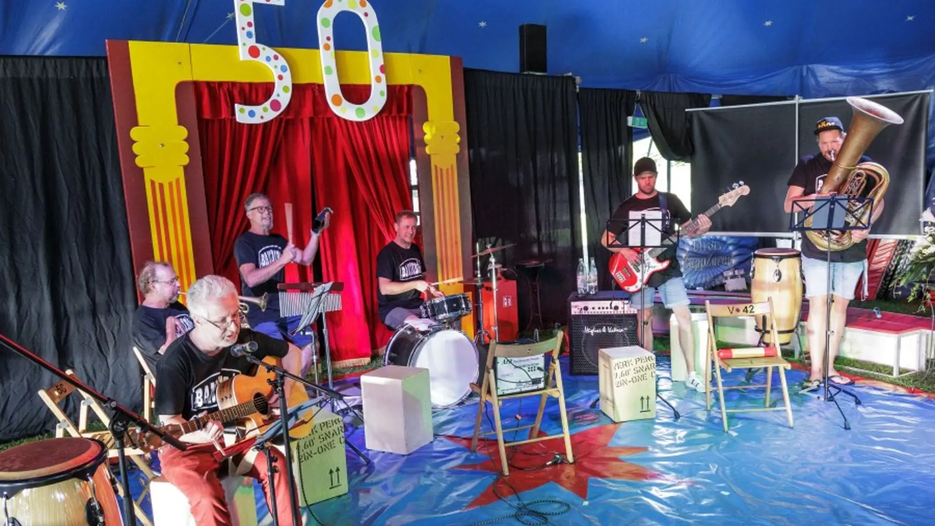Eine Band in einer Zirkusmanege. Über dem Vorhang hängt eine bunte 50.