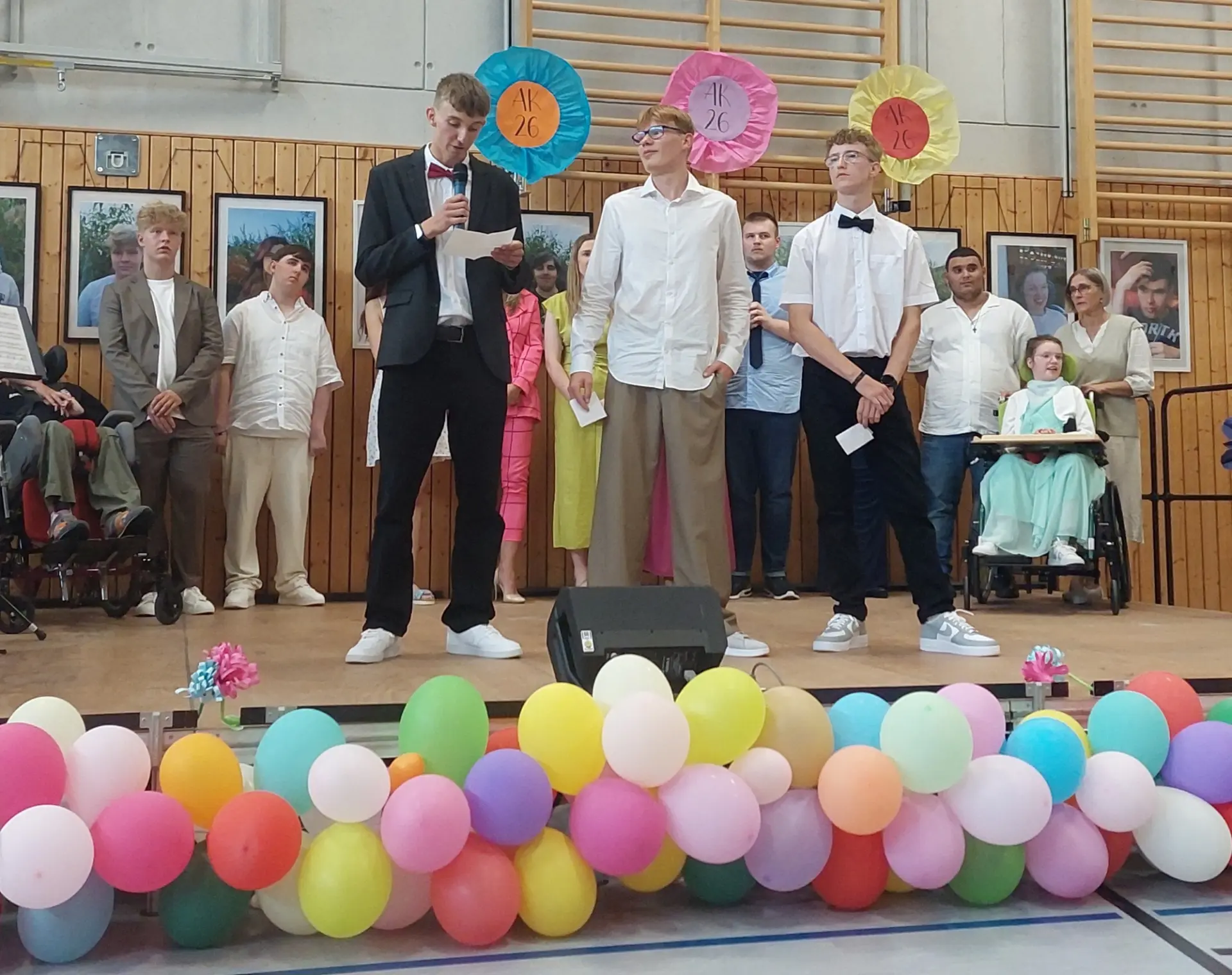 3 festlich gekleidete junge Männer stehen vor einer Gruppe auf der Bühne. Einer hält ein Mikrofon und liest von einem Stichwortzettel ab.