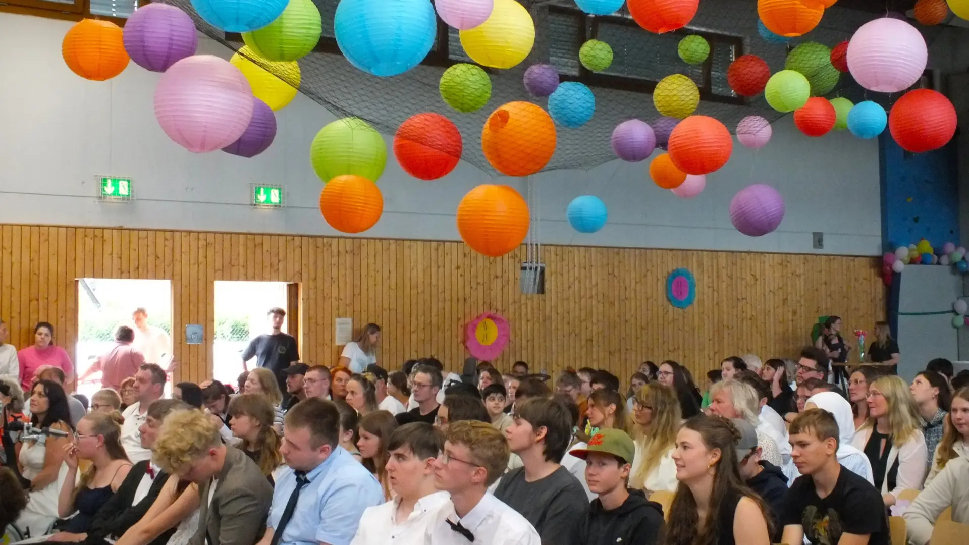 In einer mit bunten Lampions geschmückten Turnhalle sitzen viele Menschen im bestuhlten Innenraum