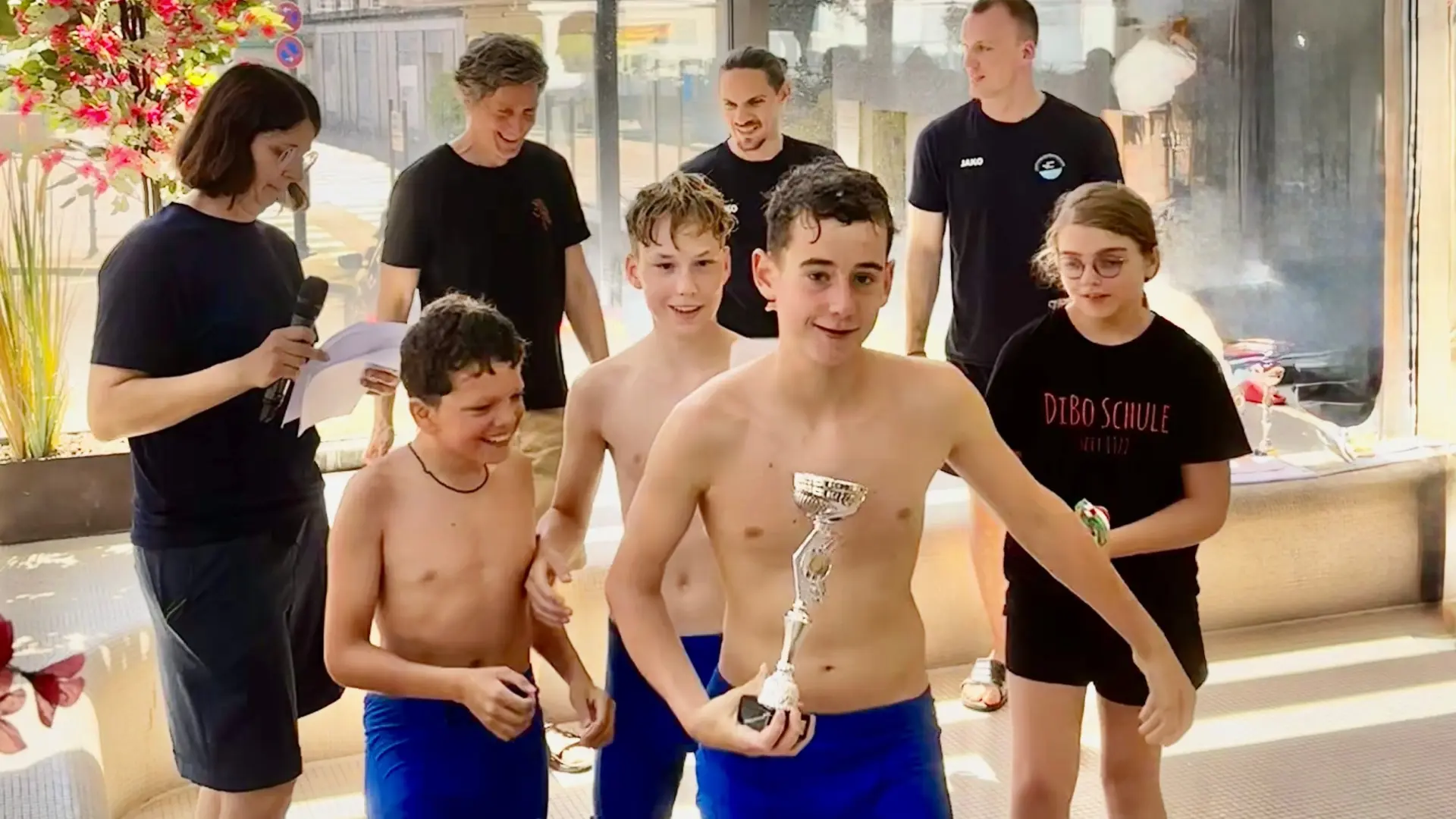 Kinder und Erwachsene im Schwimmbad am Fenster: ein Junge hält einen silbernen Pokal