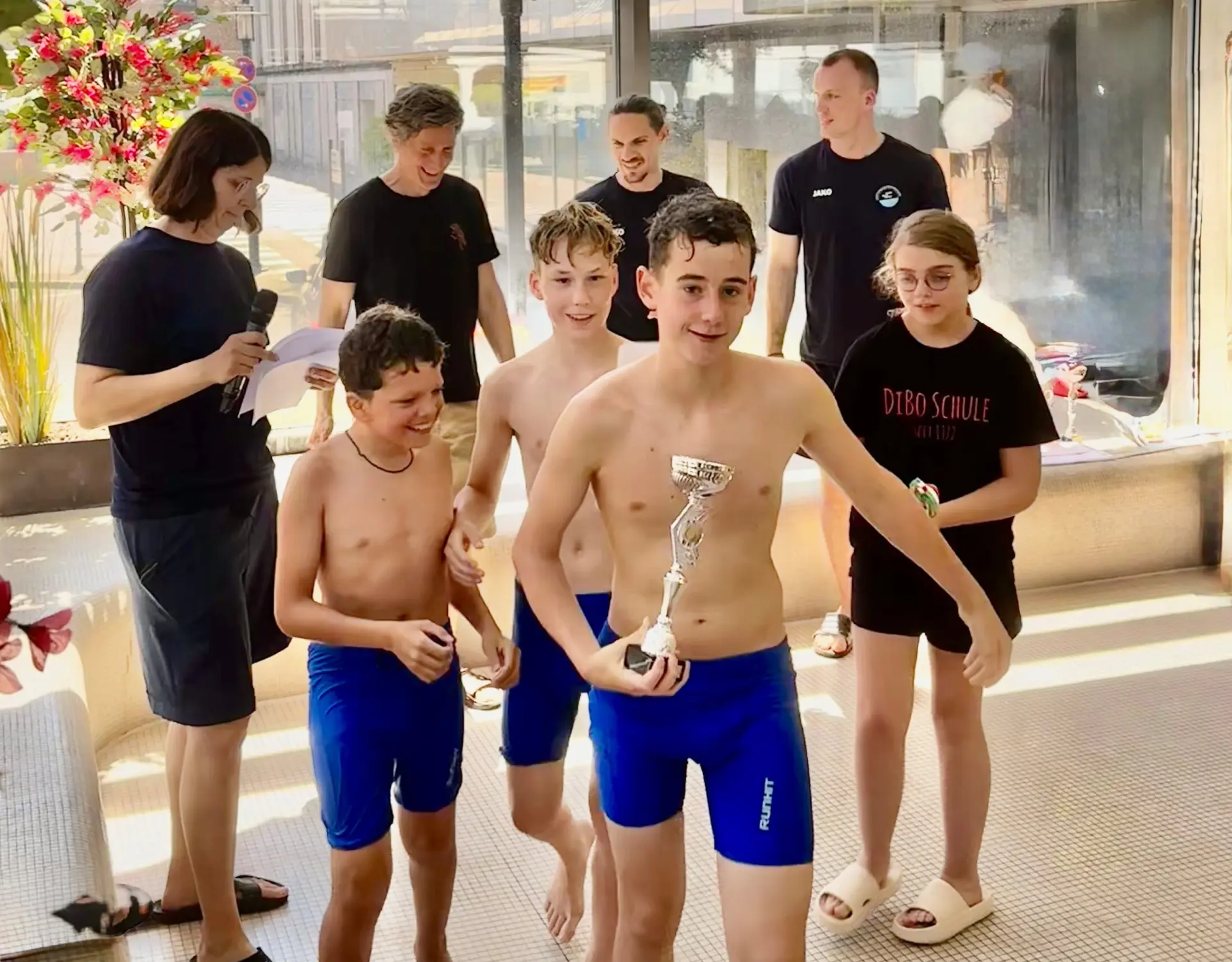 Kinder und Erwachsene im Schwimmbad am Fenster: ein Junge hält einen silbernen Pokal