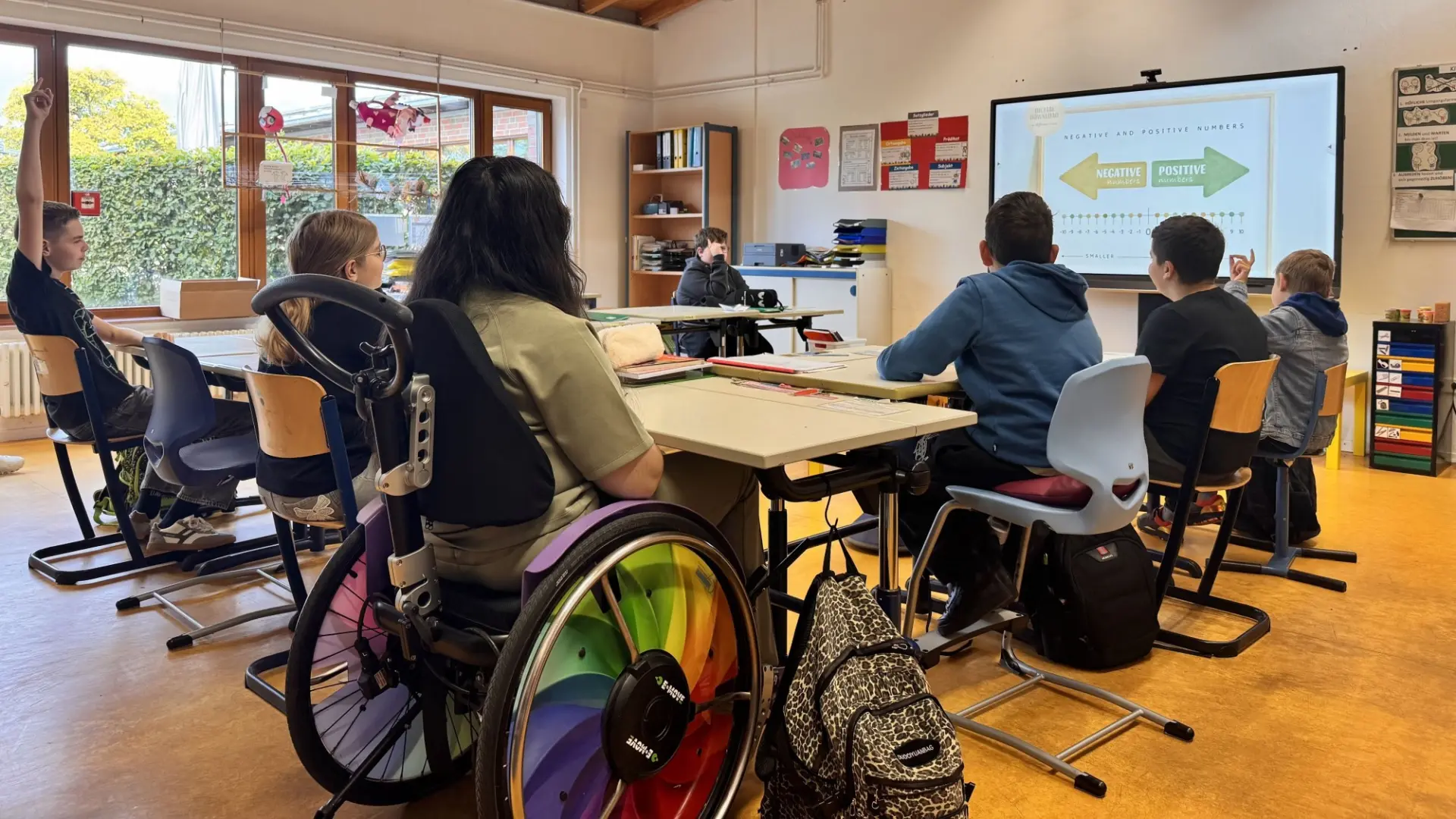 In einem freundlichen Klassenraum sitzen Schüler*innen mit und ohne Rollstuhl an Tischen vor der Tafel