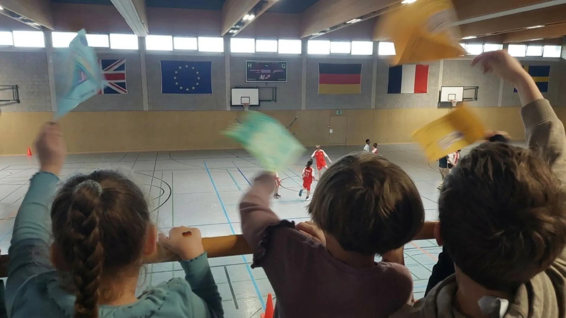Kinder jubeln mit Fähnchen Fußballspieler*innen in einer Turnhalle zu