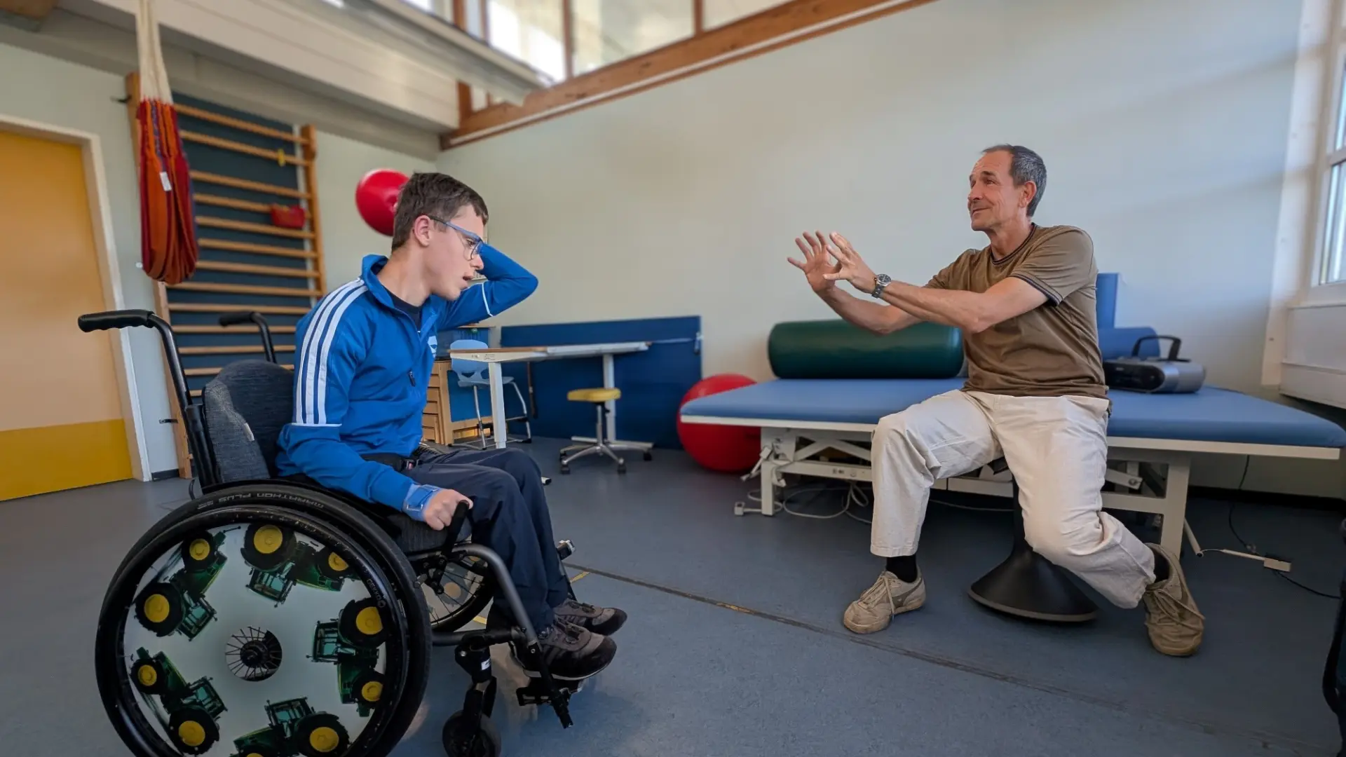 In einem großen, hellen Raum wirft ein Jugendlicher im Rollstuhl einem Mann einen Gymnastikball zuMann