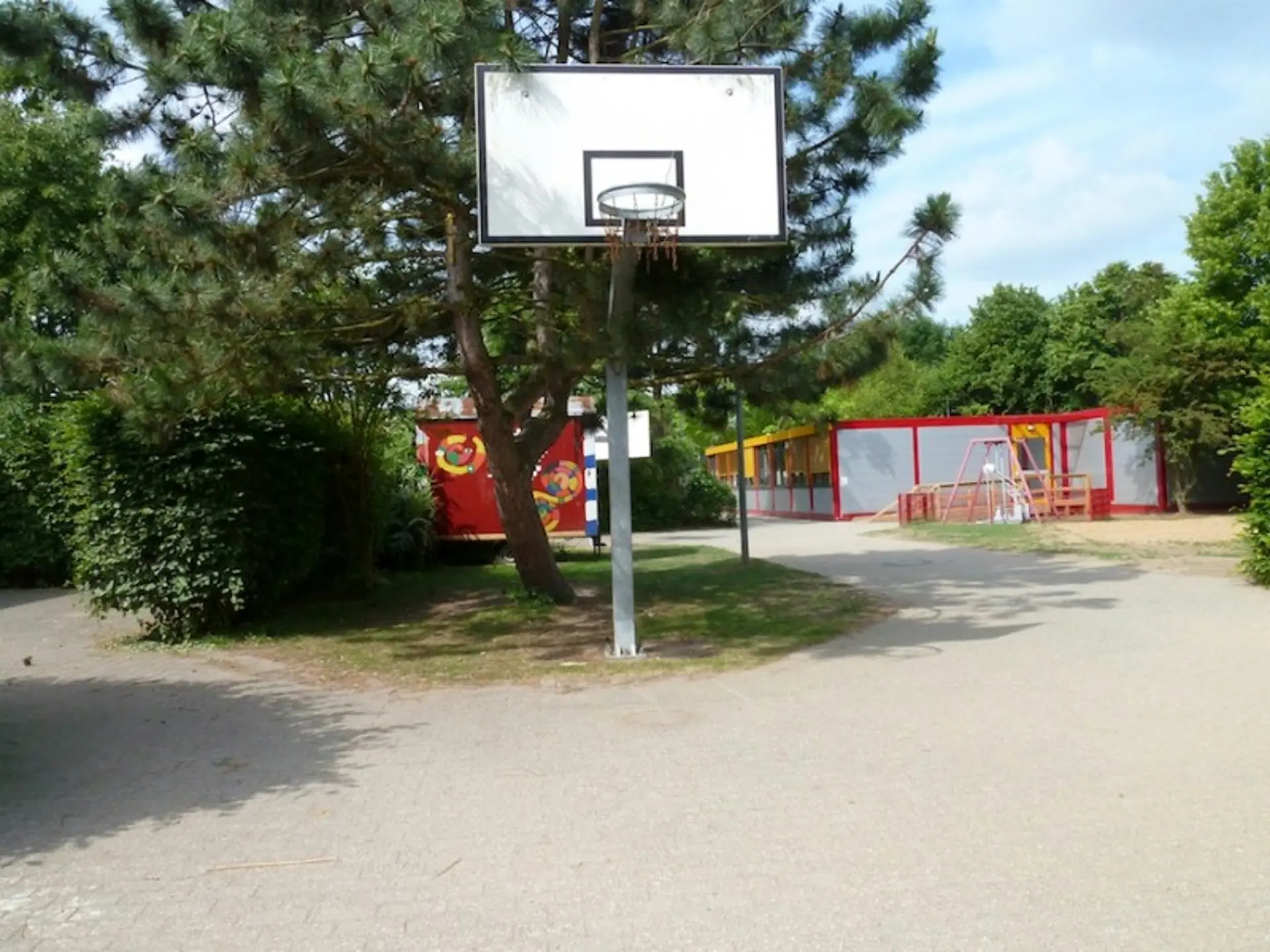 Ein Basketballkorb auf einem gepflasterten Platz im Freien, umgeben von Bäumen und Hecken im Hintergrund.