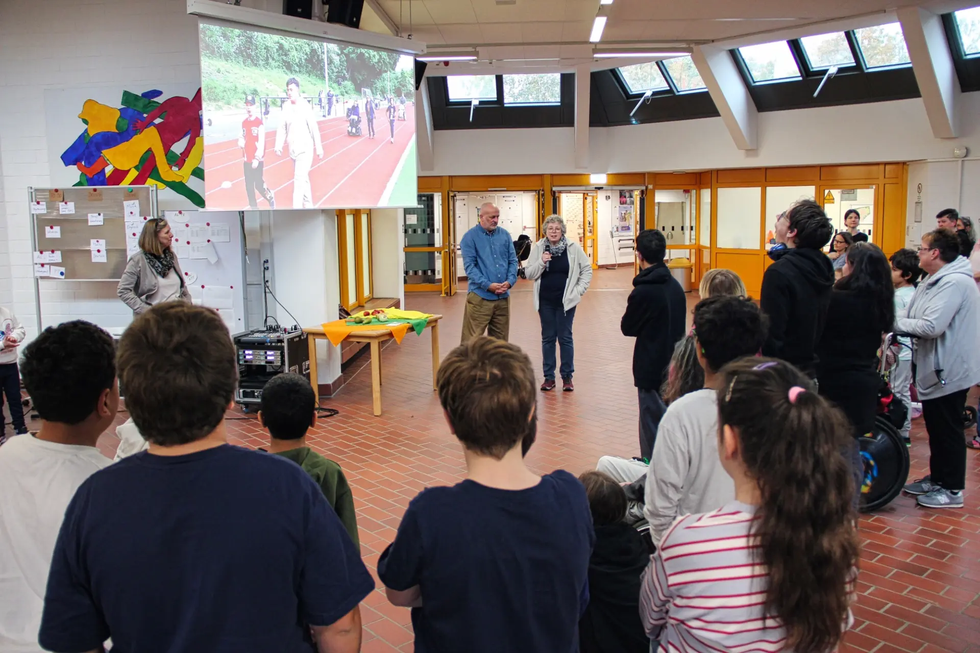 In einem großen Raum mit rotem Fußboden und gelben Fensterrahmen steht eine Gruppe Menschen im Halbkreis um eine Leinwand auf der ein Sportszene zu sehen ist.