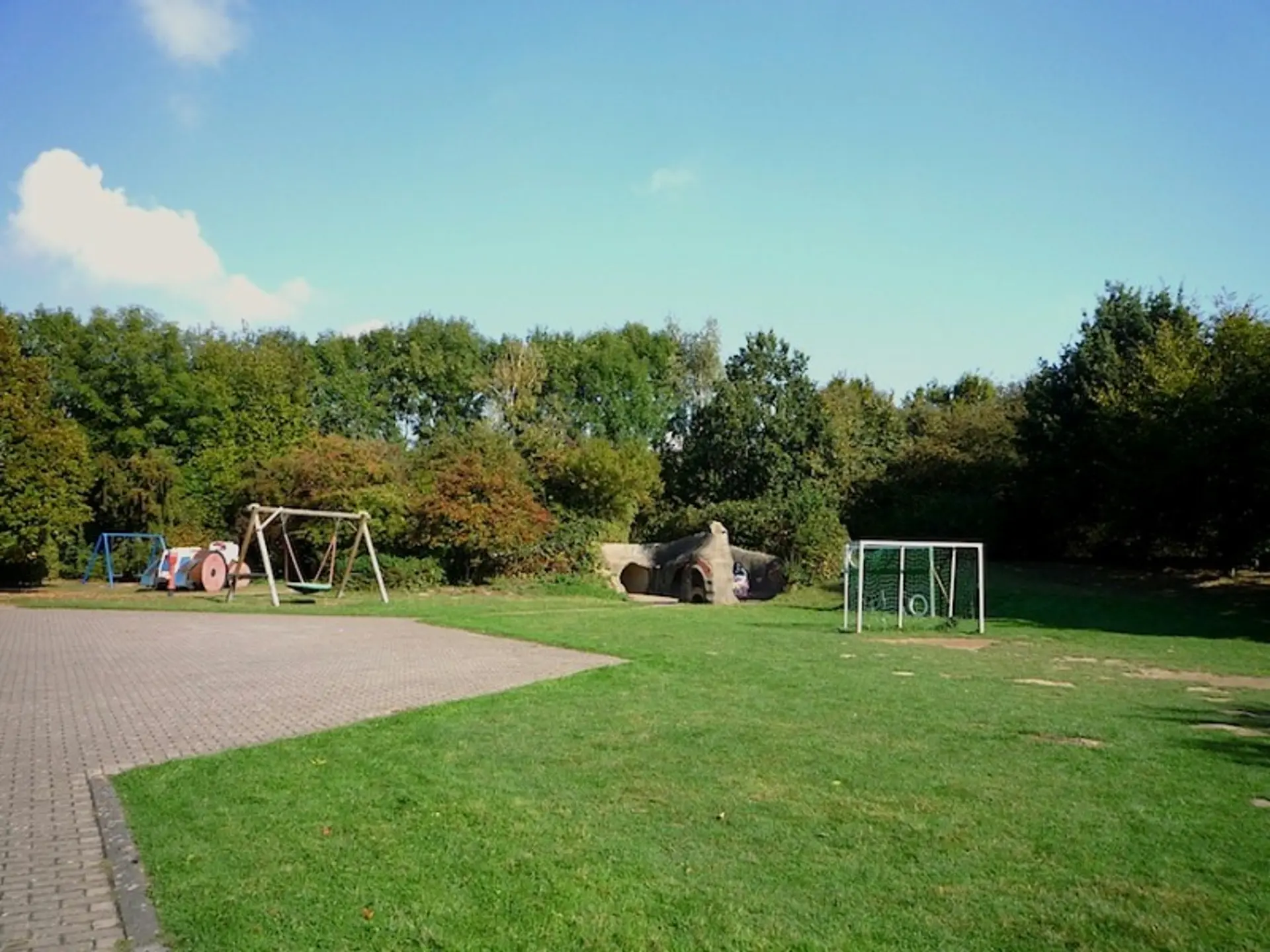 Grüne Außenanlage mit einer Nestschaukel, Fußballtoren und einem künstlichen Hügel mit Tunnel.