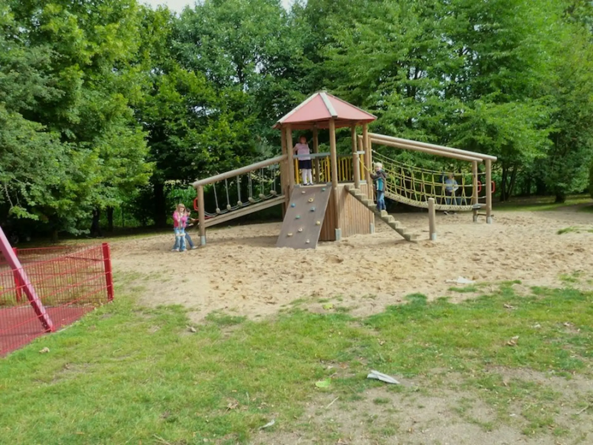 Hölzerner Spielturm mit Rutsche, Kletternetz und Brücke auf einem Sandplatz, umgeben von grünen Bäumen.