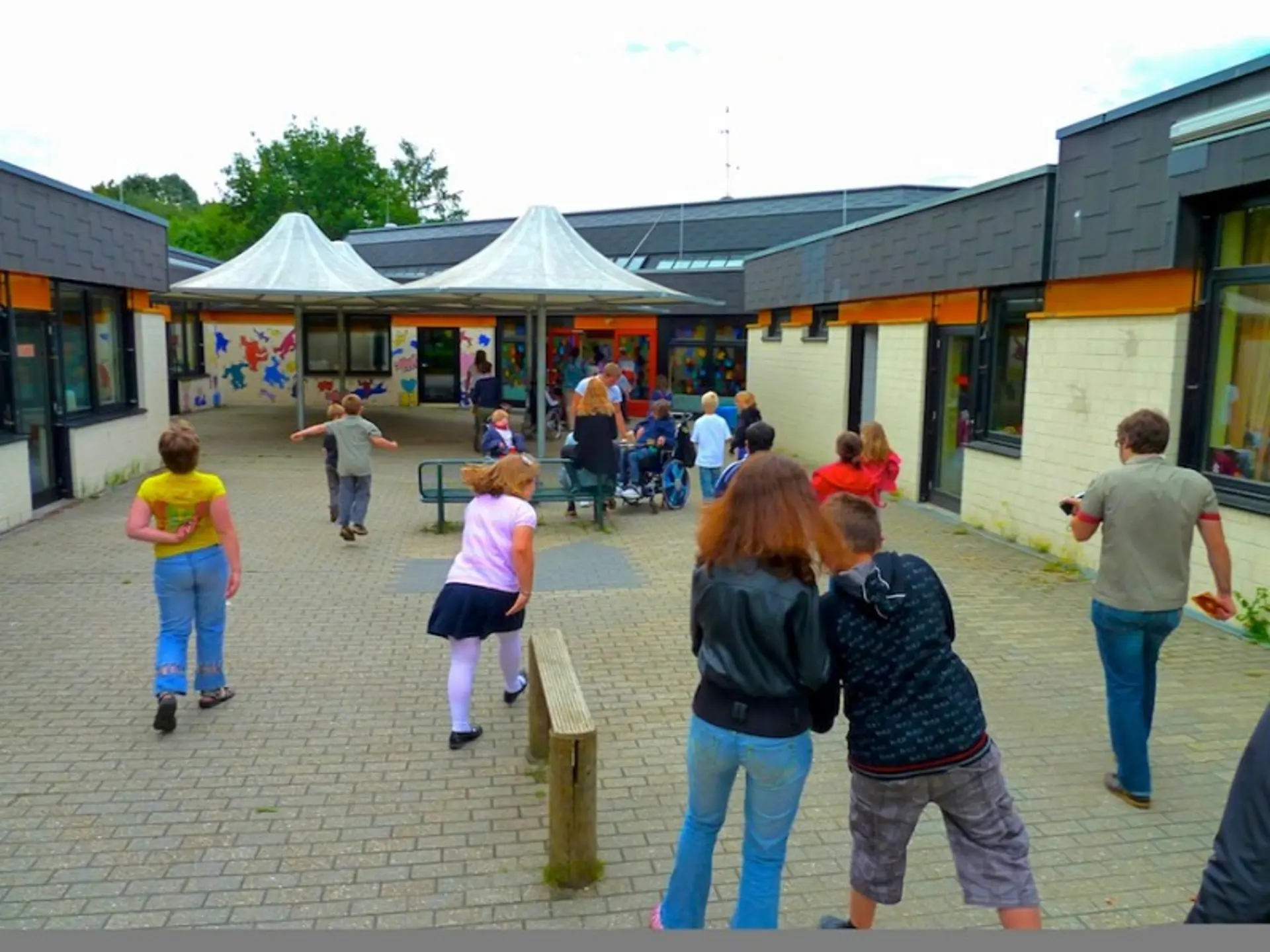 Kinder auf einem gepflasterten Innenhof einer Schule mit bunten Wandmalereien und zeltartigen Sonnendächern.
