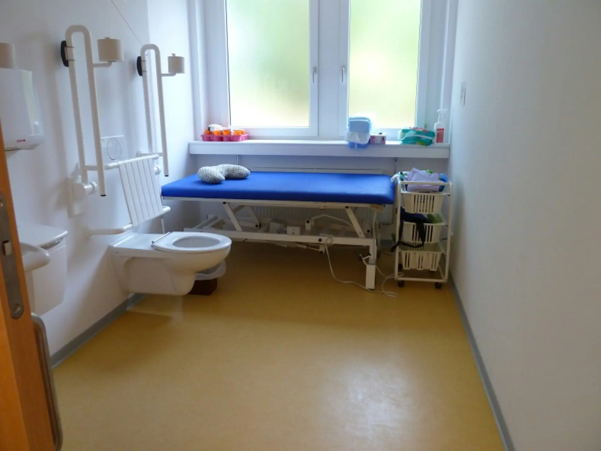 Barrierefreier Pflegeraum mit Toilette, Haltegriffen und einer blauen, höhenverstellbaren Therapieliege vor einem Fenster.