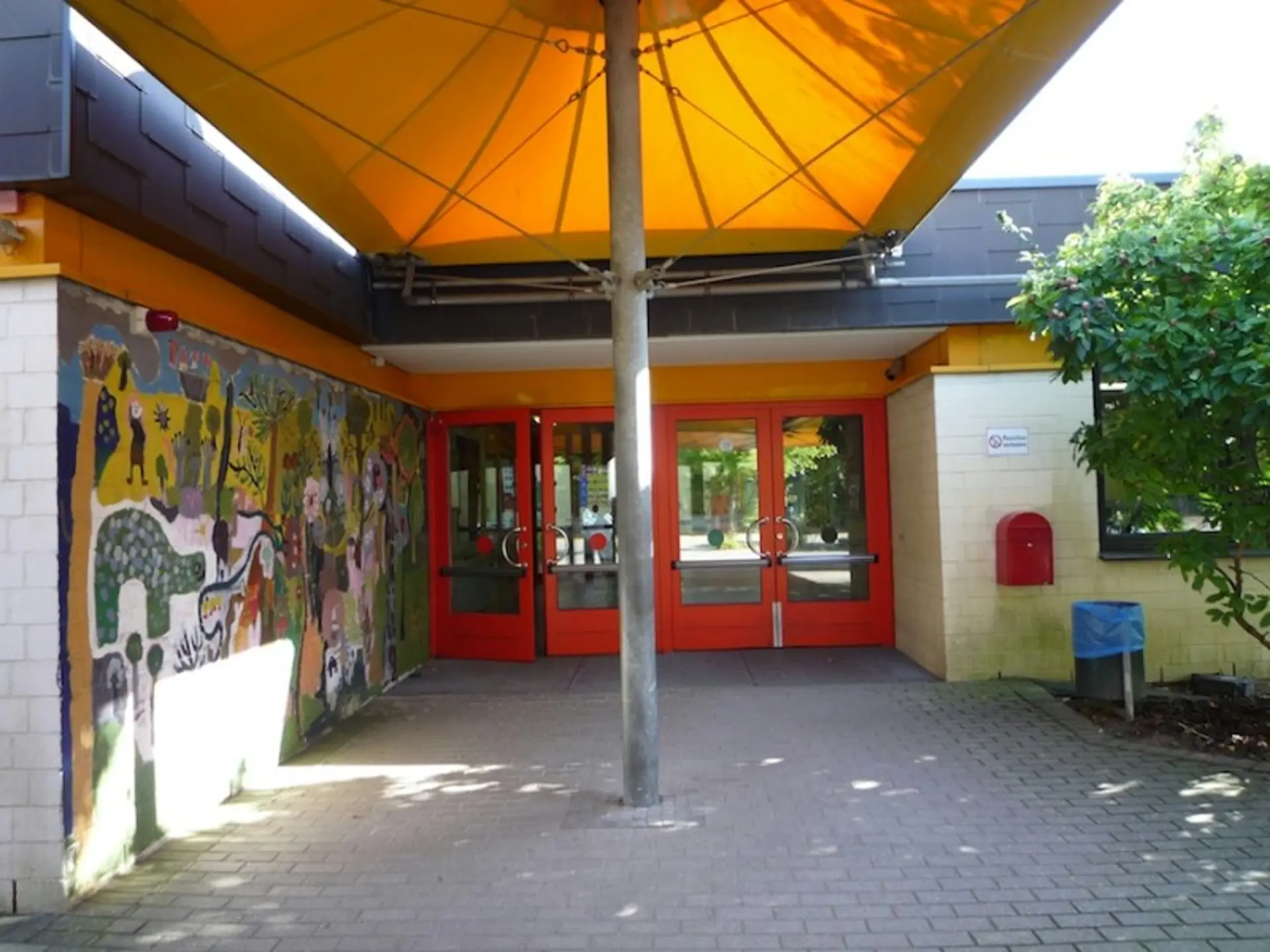 Überdachter Haupteingang mit roten Doppeltüren, einer großen Wandmalerei links und einem gelben Sonnensegel darüber.