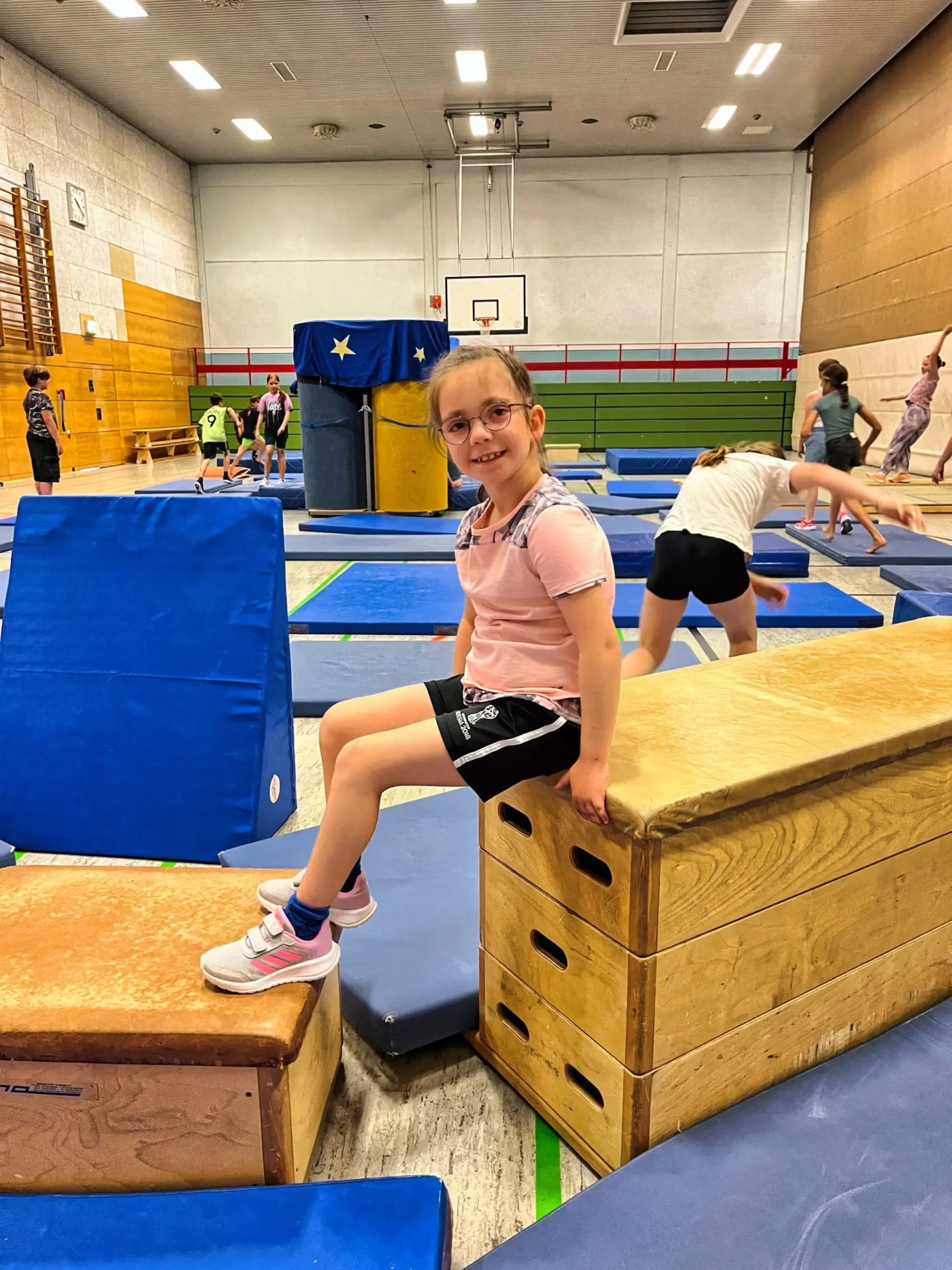 Ein Mädchen in Sportkleidung sitzt in einer Turnhalle auf einem Turnkasten.