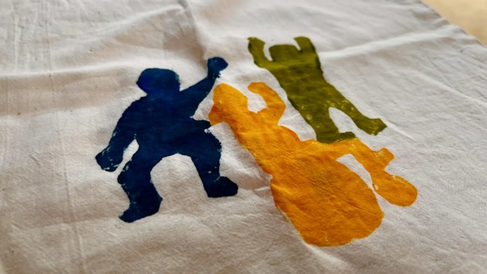 Auf ein Stück hellen Leinenstoff sind mit Plaka-Farbe drei tanzende Figuren gemalt: in blau, in orange und in hellgrün. Die orangene Figur sitzt in einem Rollstuhl, der ebenfalls orange ist.