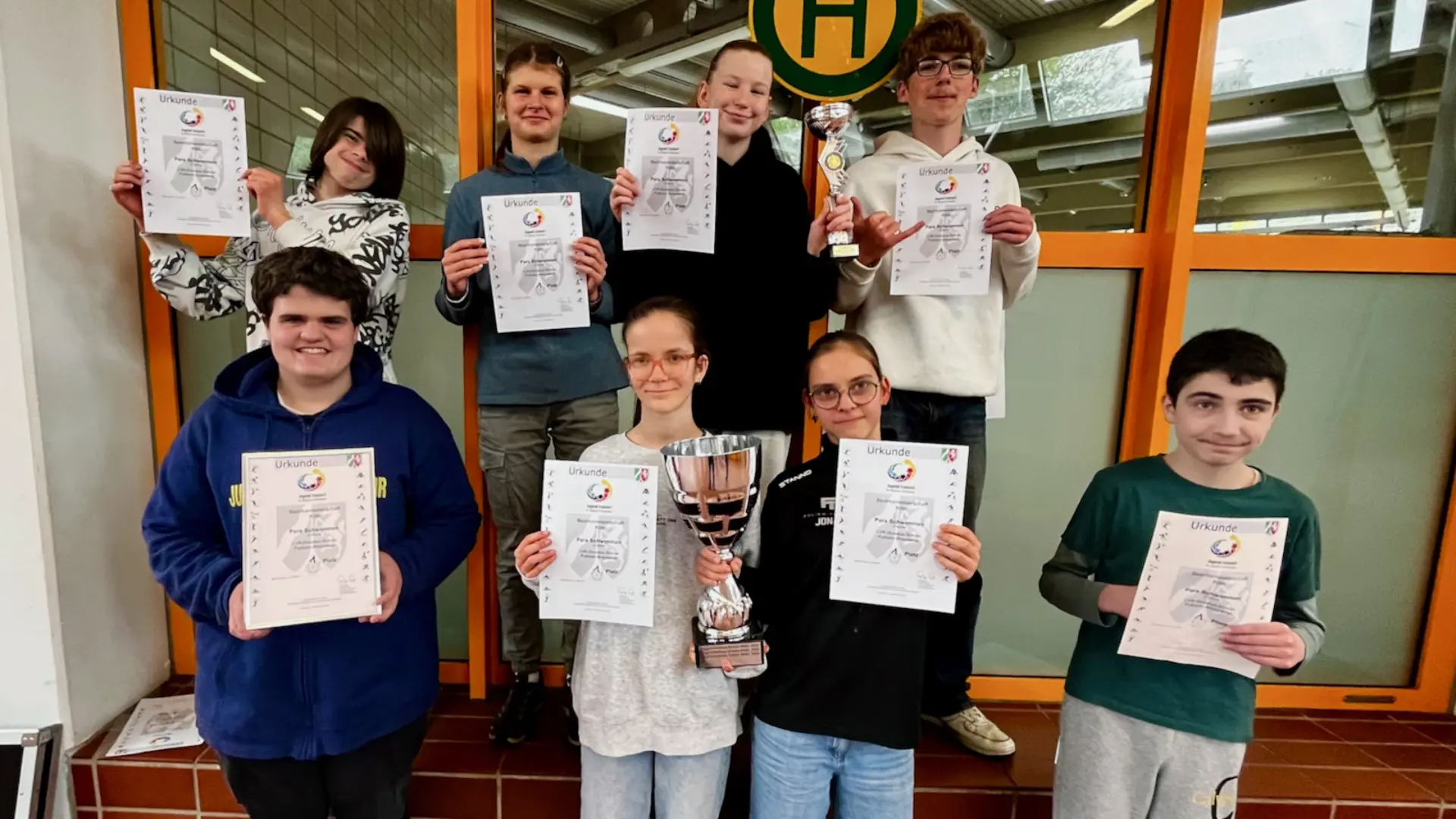 8 Jugendliche schauen glücklich in die Kamera. Sie halten ihre Urkunden ins Bild. Ein Mädchen hält einen großen silbernen Pokal, ein anderes einen kleinen silbernen Pokal in der Hand.
