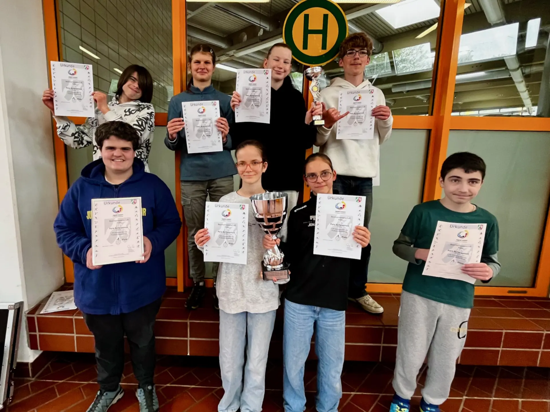8 Jugendliche schauen glücklich in die KAmerea. Sie halten Urkuinde ins Bild. Ein Mädchen hält einen großen silbernen Pokal, ein anderes einen kleinen silbernen Pokal in der Hand.