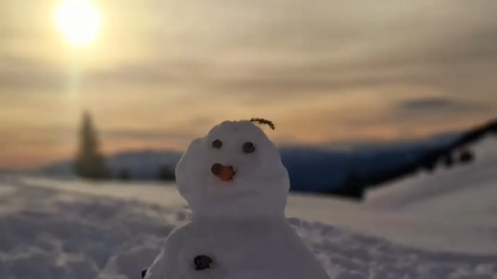 Ein Schneemann vor einer schneebedeckten Bergkulisse. Im Hintergrund geht golden die Sonne unter.