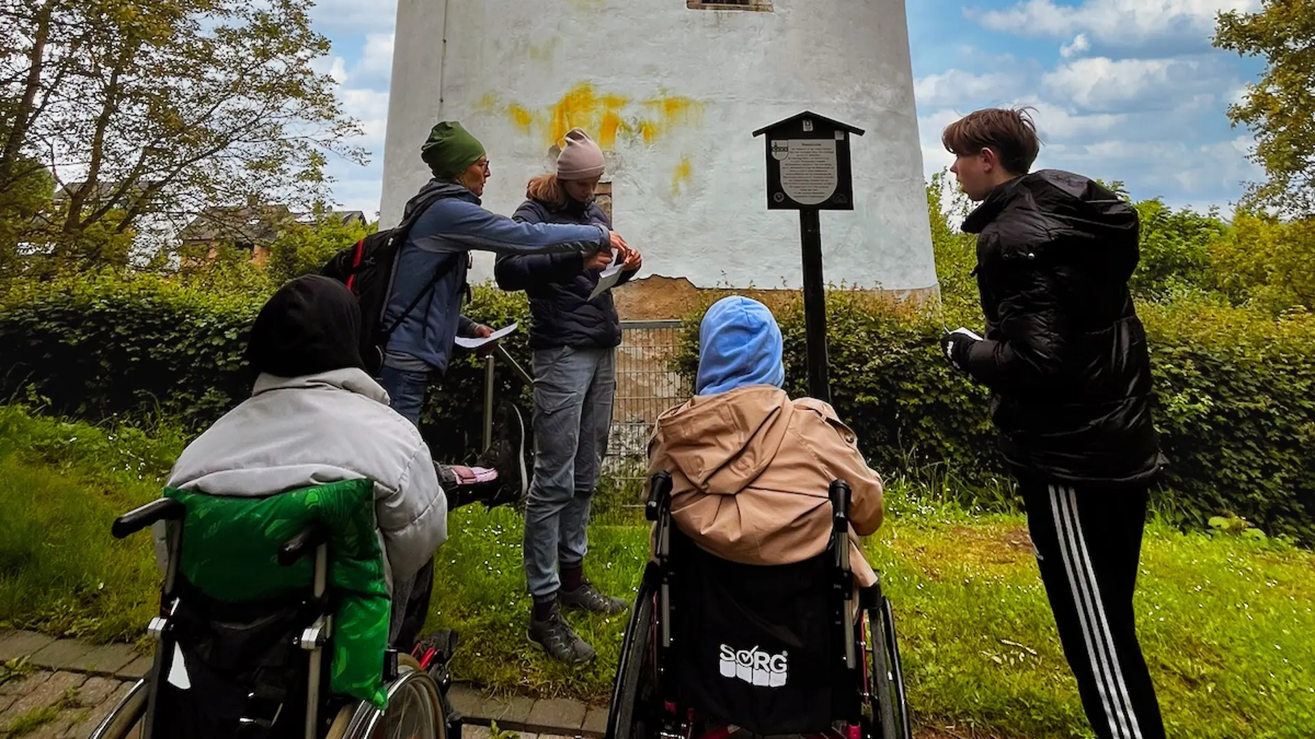 Vier Jugendliche, davon zwei im Rollstuhl vor einem runden, weißen Turm im Grünen. Der Himmel ist strahlend blau mit kleinen weißen Wolken.