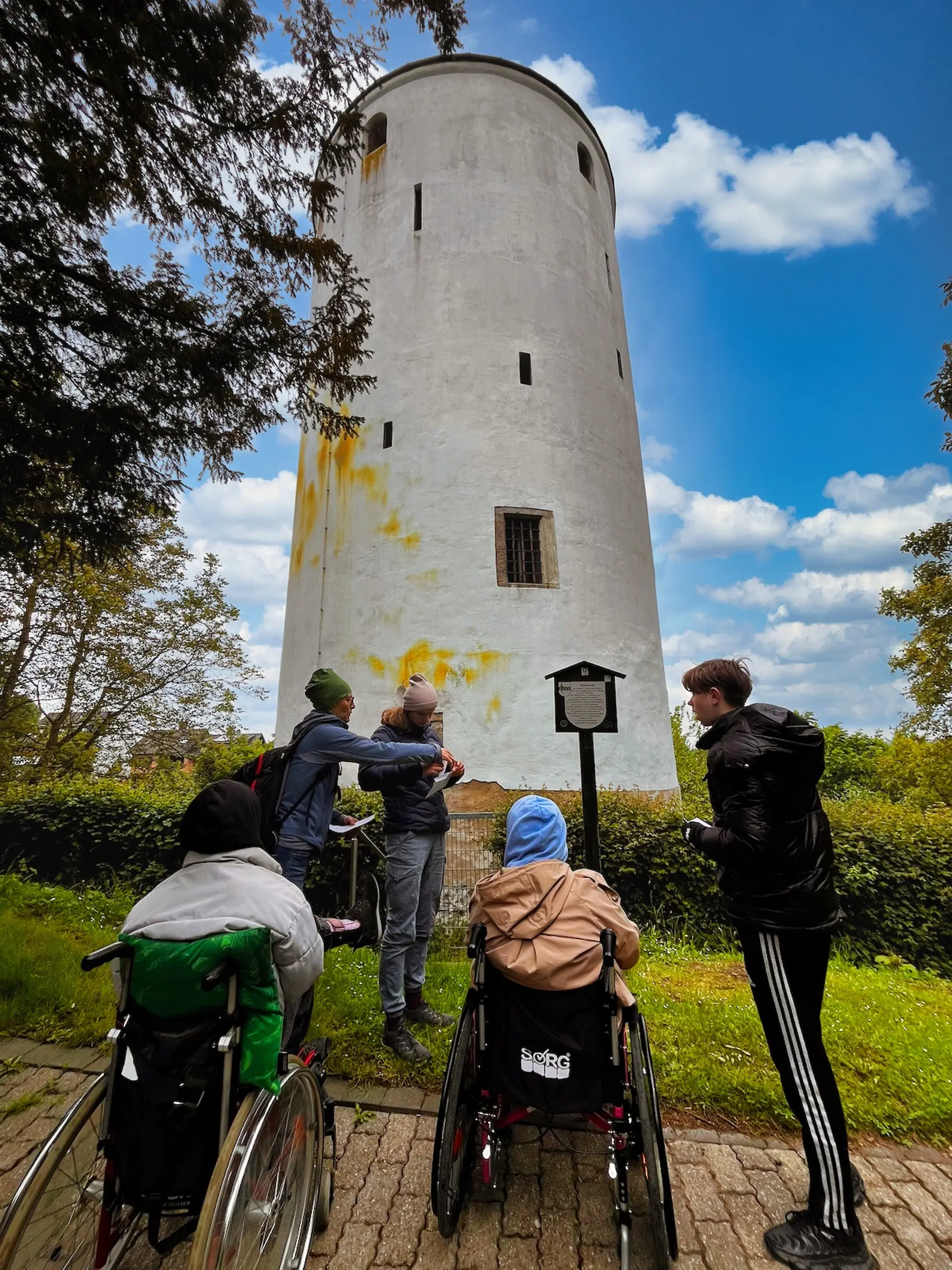 Vier Jugendliche, davon zwei im Rollstuhl vor einem runden, weißen Turm im Grünen. Der Himmel ist strahlend blau mit kleinen weißen Wolken.
