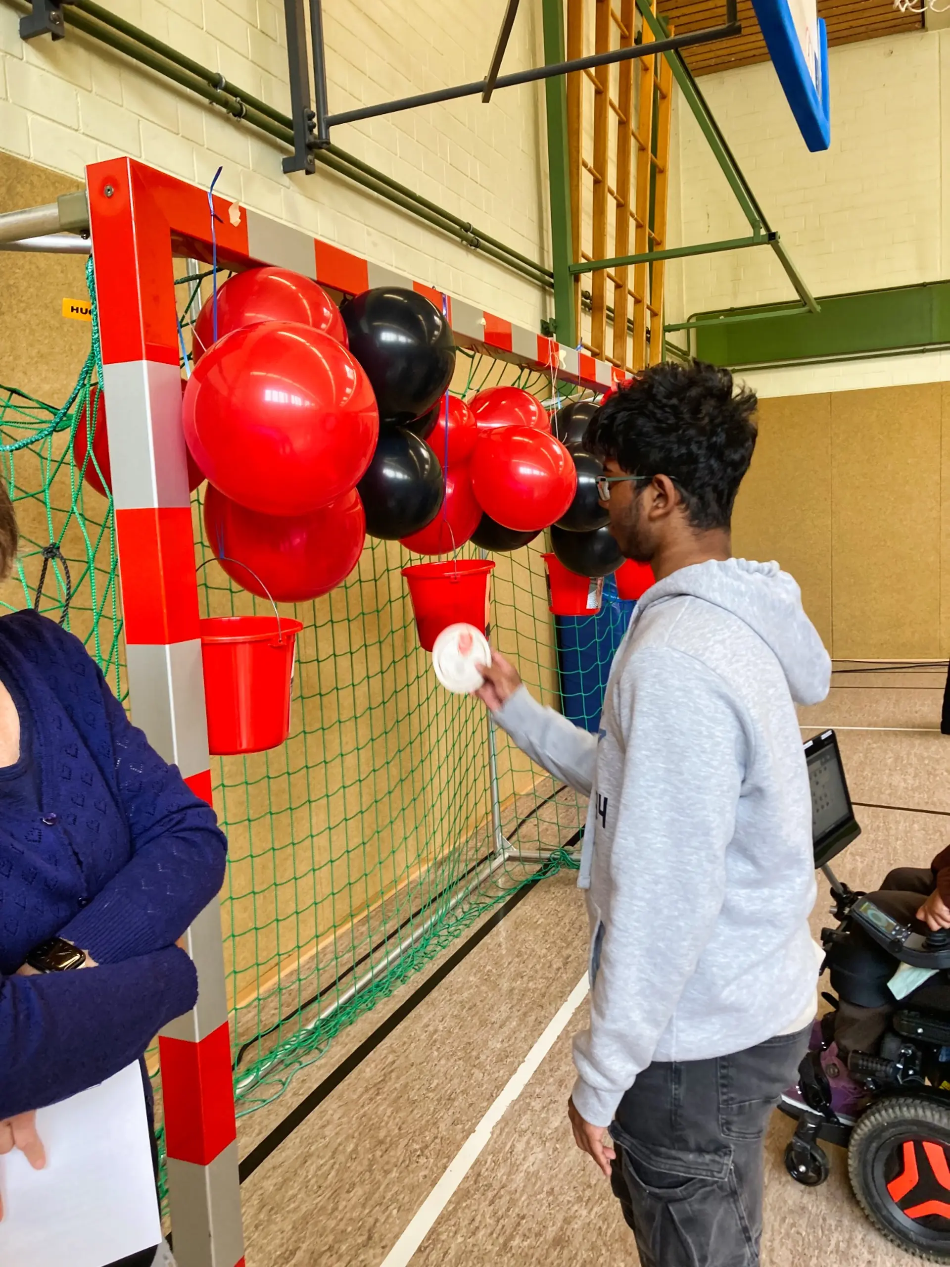 In einer Turnalle sind an einem Fußballtor rote und schwarze Ballons befestigt und vier rote Eimer. Ein Jugendlicher hält einen Bierdeckel in der Hand. Erscheint zu überlegen, in welchen Eimer er ihn legen soll.
