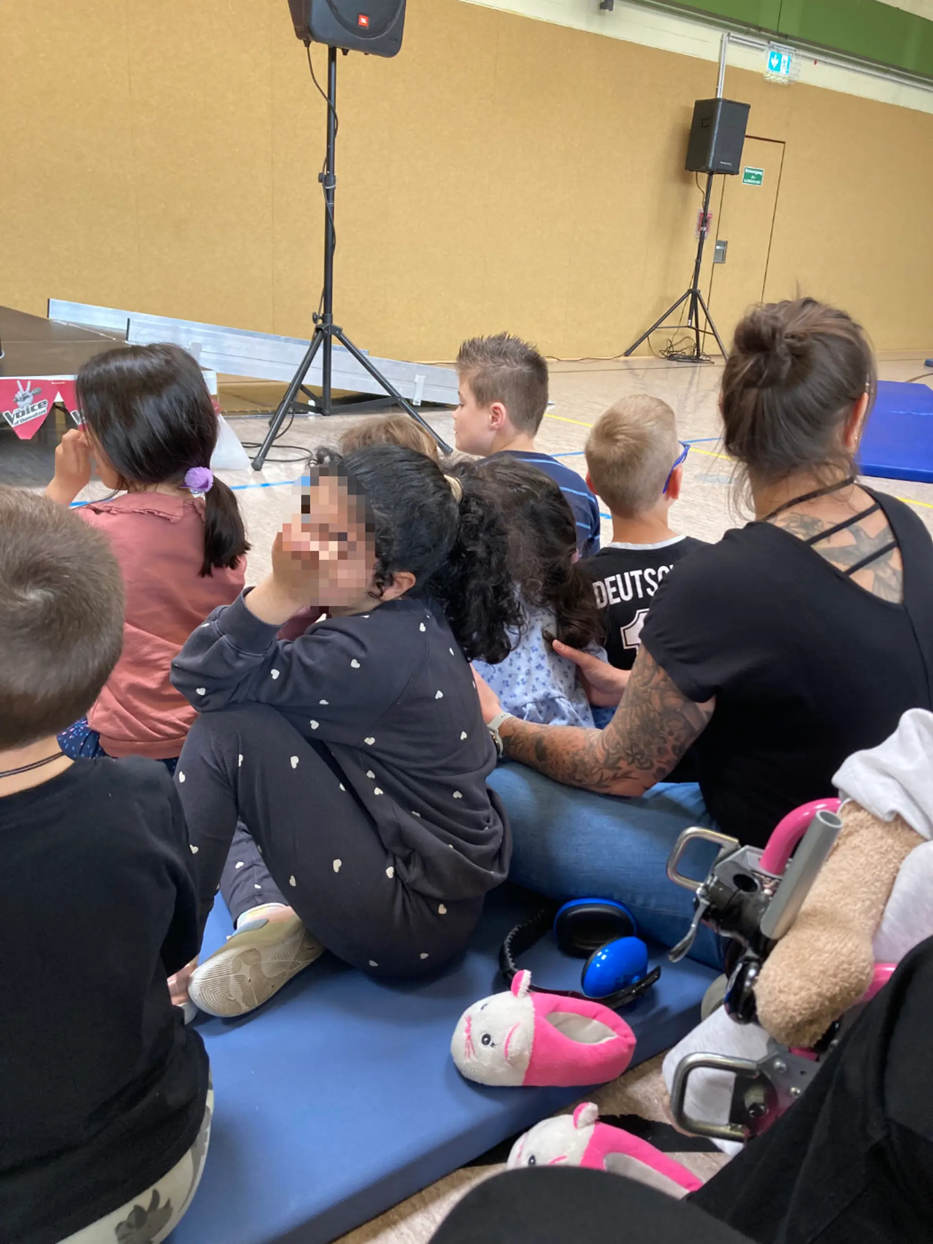 Auf blauen Turnmatten sitzen Kinder und Erwachsen in einer Turnhalle und schauen auf eine Bühne. Neben der Bühne stehen große Lautsprecher. Auf die Bühne führt eine Rampe aus Metall.