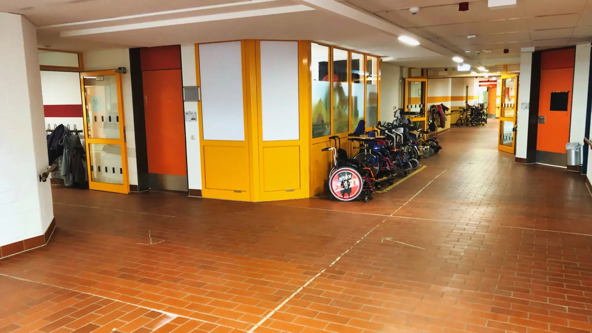 Ein heller Schulflur mit rotem Fliesenboden, Garderobe links und gelber Trennwand. Rechts sind mehrere Rollstühle, Gehhilfen und Rollatoren geparkt, darunter ein Rollstuhl mit bedruckter Radkappe.
