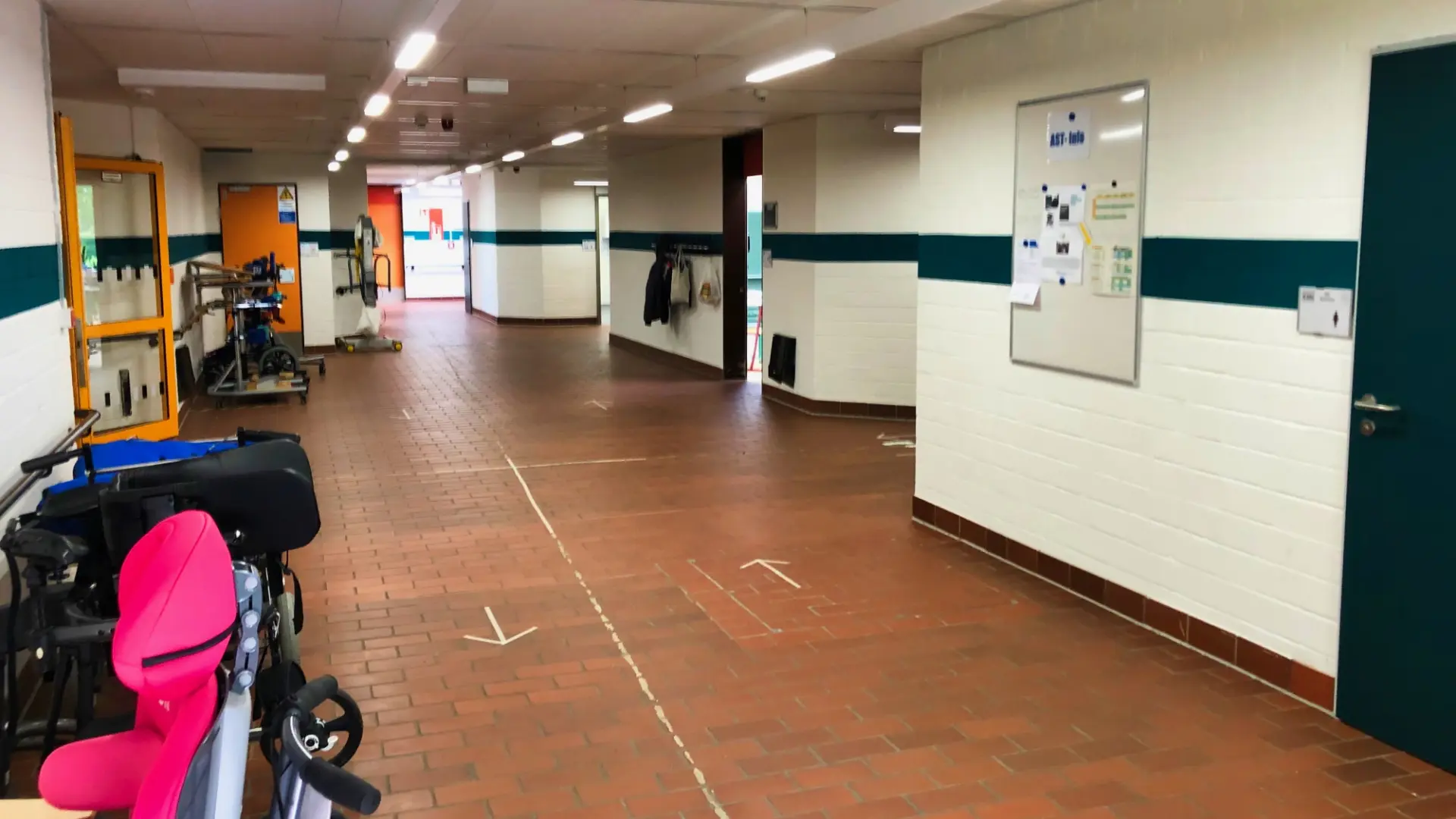 Ein weiter Schulflur mit Backsteinboden und grünen Wandstreifen. Vorne links steht ein pinker Spezialsitz, im Hintergrund parken Rollstühle. Rechts hängt ein Infobrett an der weißen Wand.