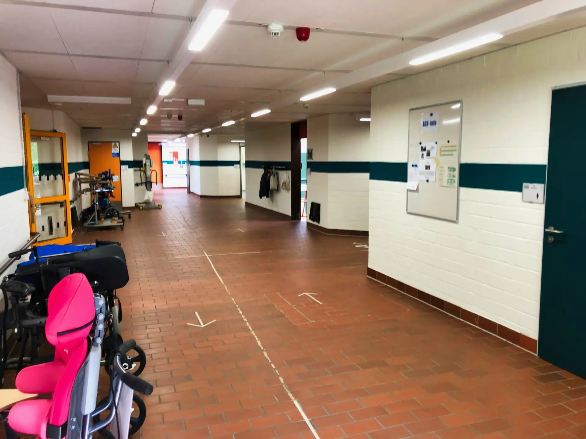 Ein weiter Schulflur mit Backsteinboden und grünen Wandstreifen. Vorne links steht ein pinker Spezialsitz, im Hintergrund parken Rollstühle. Rechts hängt ein Infobrett an der weißen Wand.