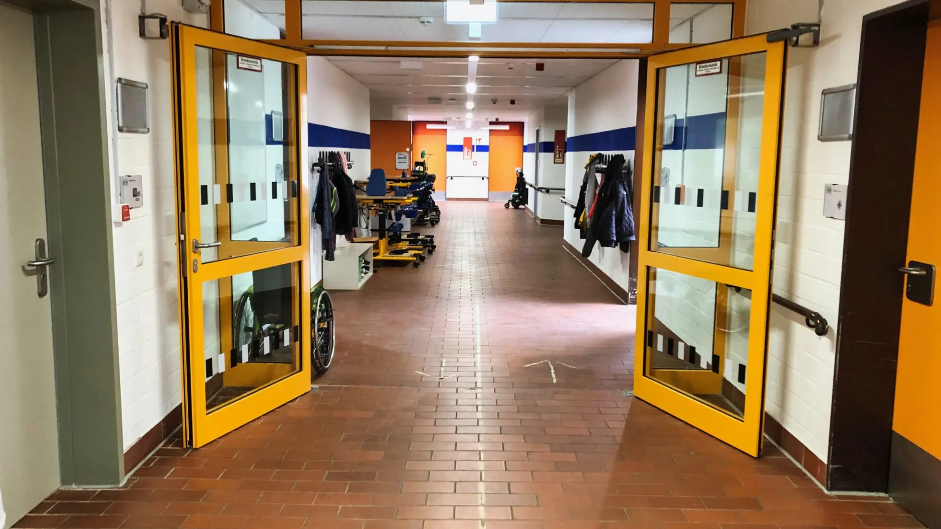 Ein heller Schulflur mit rotbraunem Fliesenboden, blauen Wandstreifen und geöffneten gelben Flügeltüren. Links und rechts hängen Jacken an Haken, dahinter stehen Rollstühle und Gehhilfen.