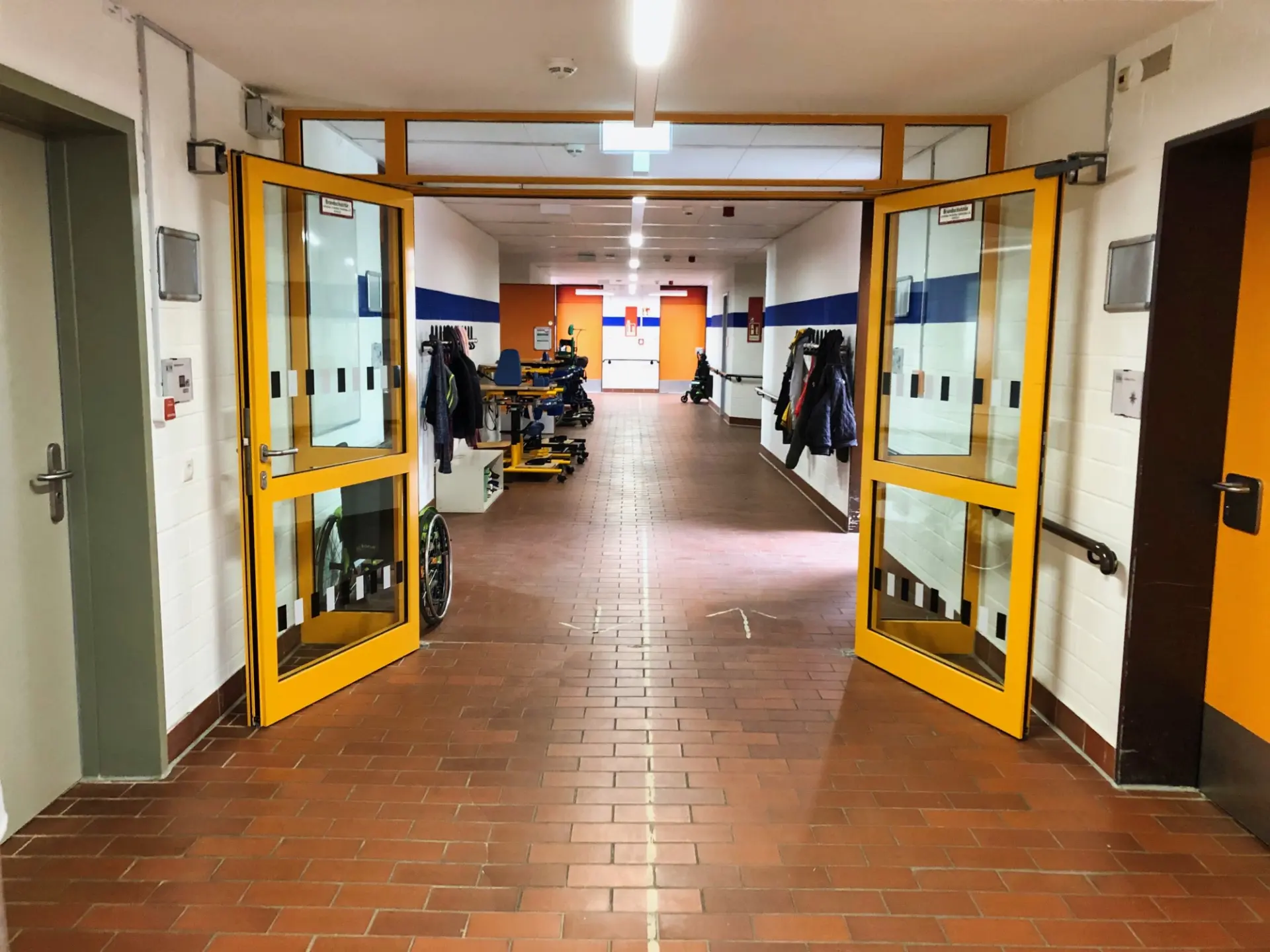 Ein heller Schulflur mit rotbraunem Fliesenboden, blauen Wandstreifen und geöffneten gelben Flügeltüren. Links und rechts hängen Jacken an Haken, dahinter stehen Rollstühle und Gehhilfen.