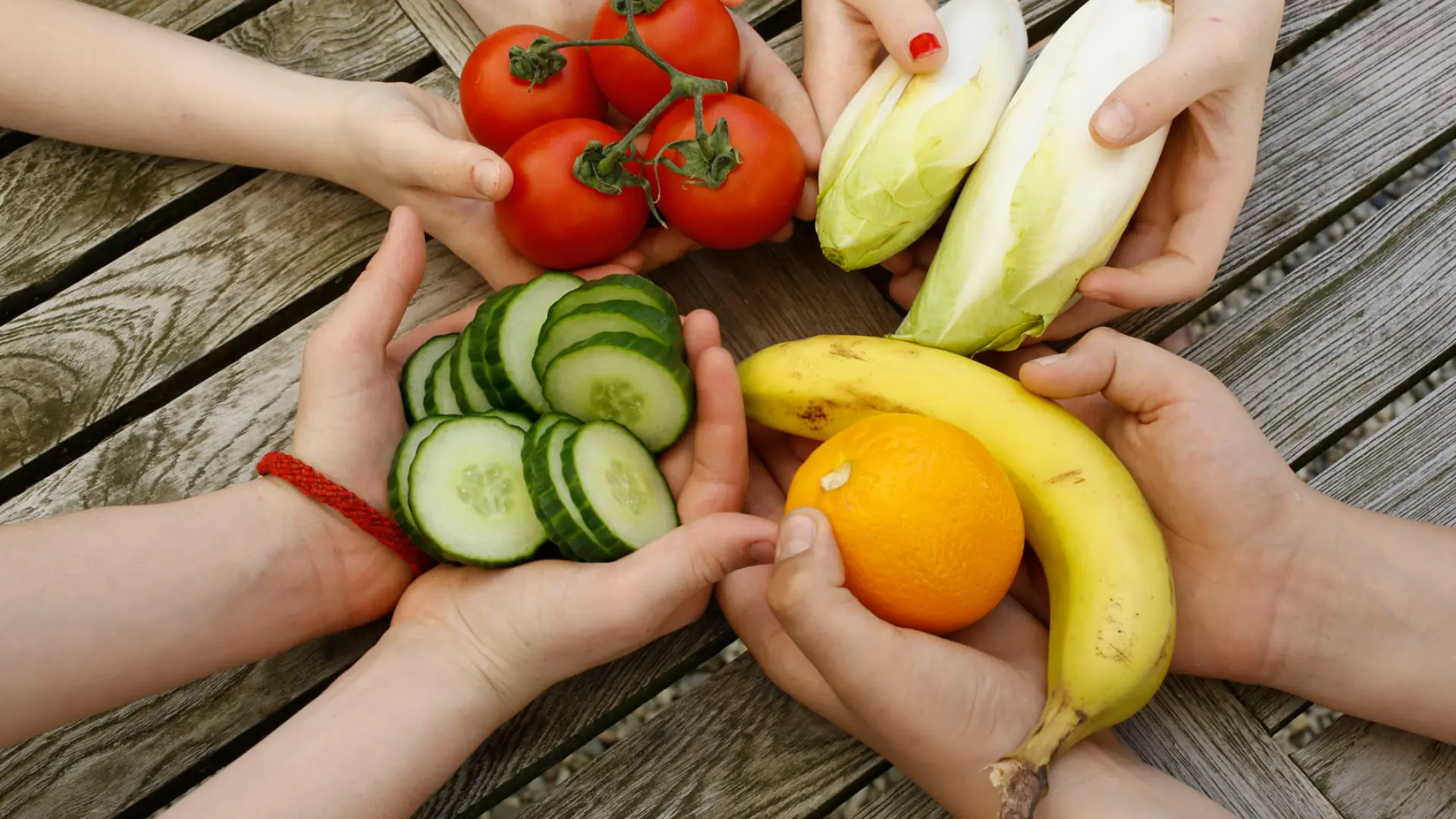 Vier Paar Kinderhände halten im Kreis über einem Holztisch frisches Obst und Gemüse: Tomaten am Strauch, Chicorée, Gurkenscheiben sowie eine Banane und eine Orange.