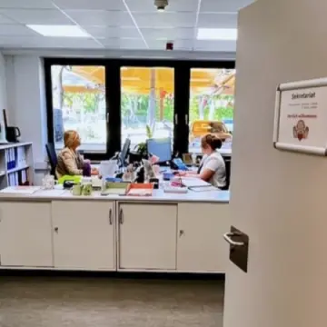 Blick in ein Büro. An der Tür ein Schild "Sekretariat". Im Raum sitzen zwei Personen an Schreibtischen.