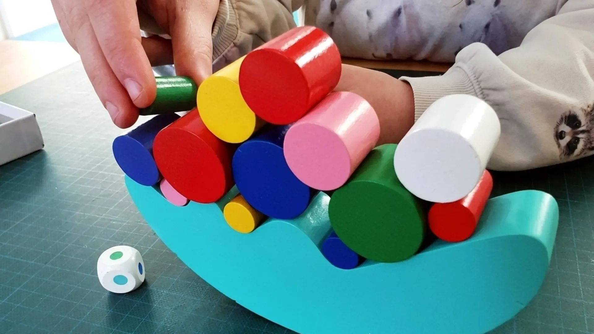 Eine Kinderhand stapelt konzentriert bunte, zylindrische Holzklötze auf ein türkisfarbenes, wippenartiges Spielbrett. Ein kleiner Würfel liegt auf der dunkelgrünen Unterlage.
