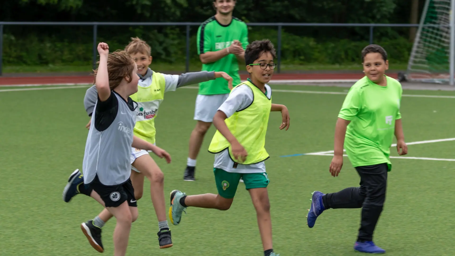 Vier Jungs in Bewegung auf einem Kunstrasenfeld bei Fußballspielen, Sie freuen sich, als hätten sie gerade ein Tor geschossen. Im Hintergrund klatscht ein Erwachsener, der ebenfalls in Sportkleidung auf dem Feld steht.