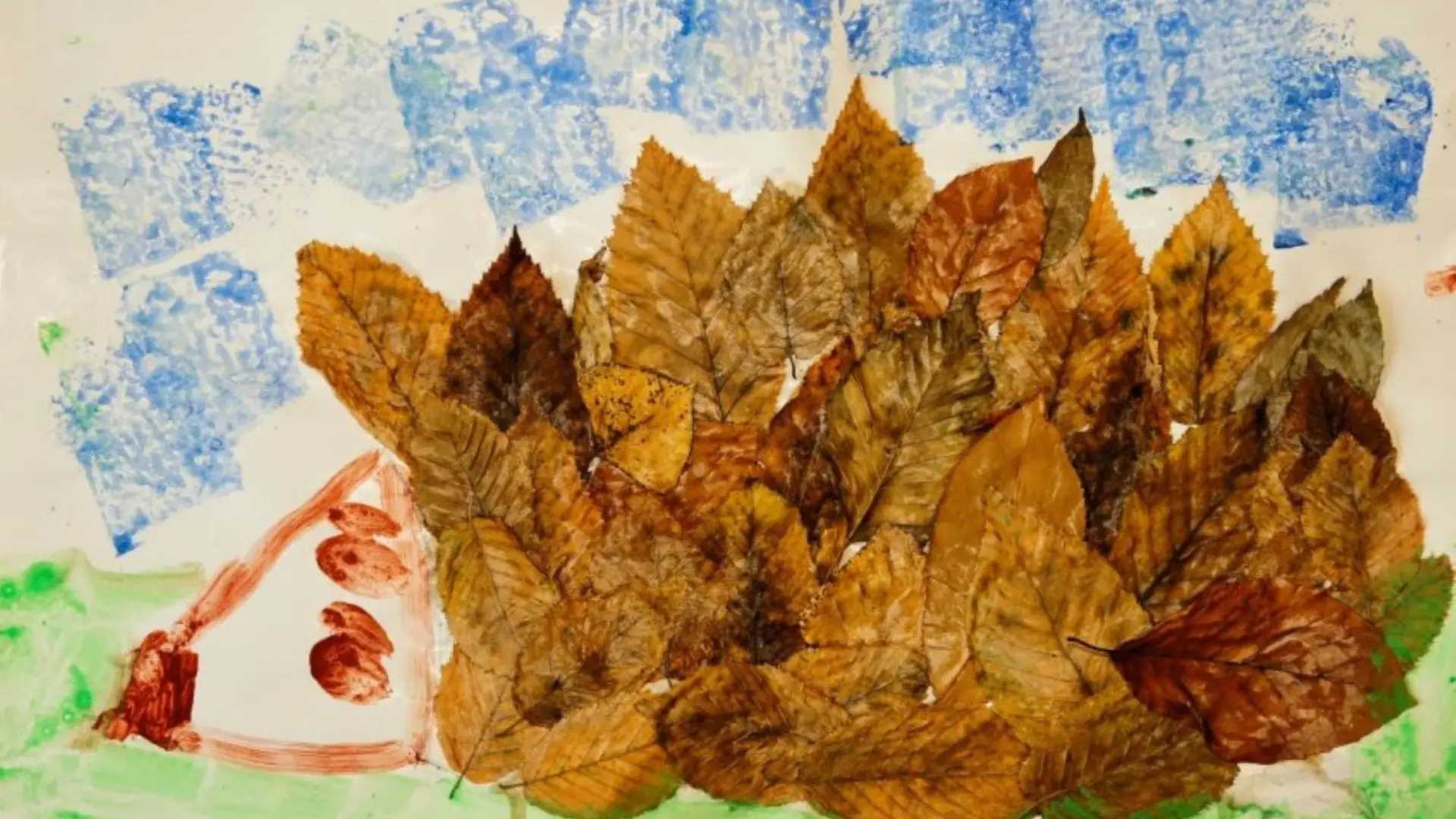 Bild eines Igels, gebastelt aus echten, braunen Herbstblättern, die seine Stacheln bilden. Das Gesicht ist aufgemalt, oben sind blaue Farbabdrücke zu sehen und unten grünes Gras.