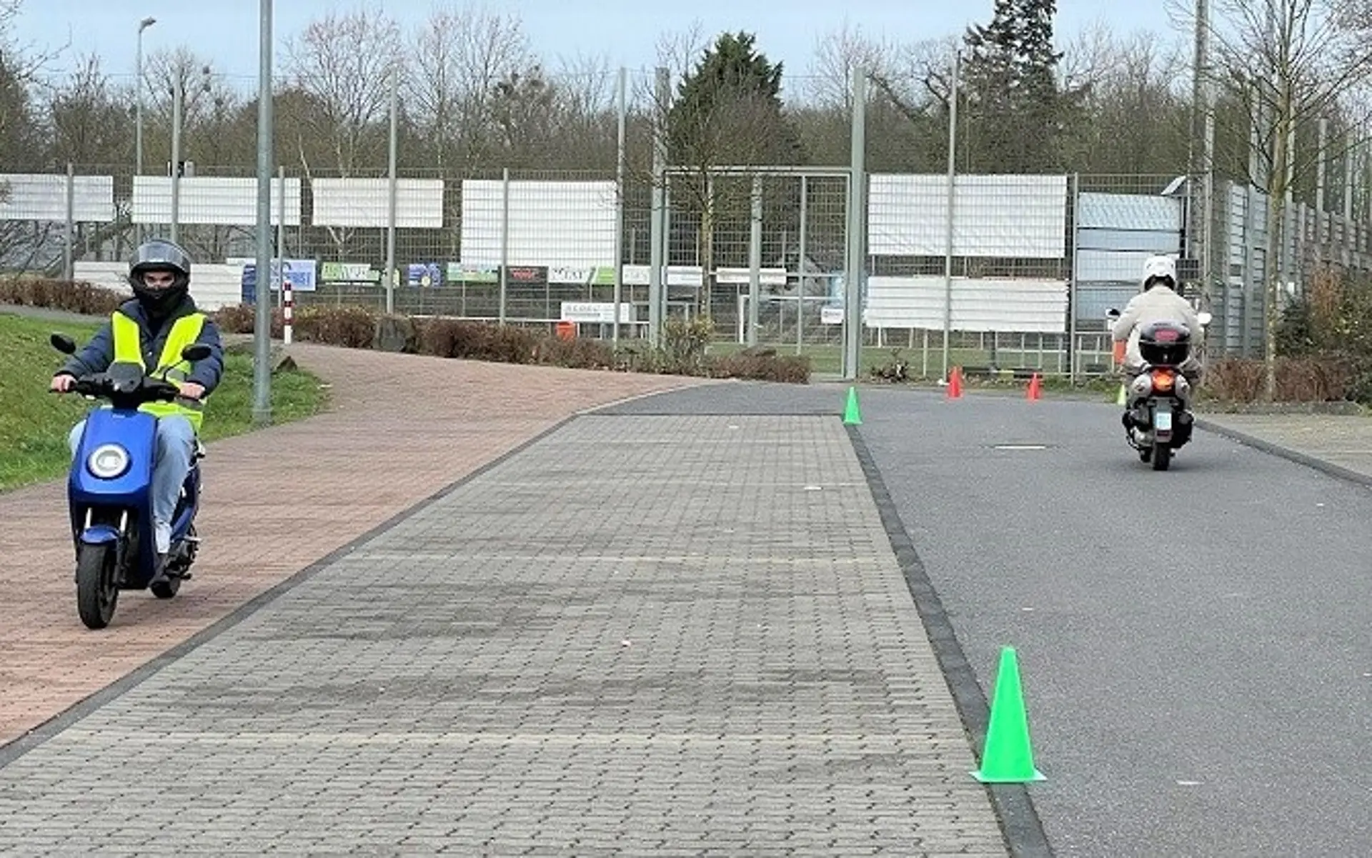 Zwei Schüler absolvieren ein praktisches Mofa-Fahrtraining auf einem Übungsplatz. Ein Schüler trägt eine Warnweste und einen Helm, während er mit einem Mofa einen Parcours mit grünen Pylonen durchfährt.