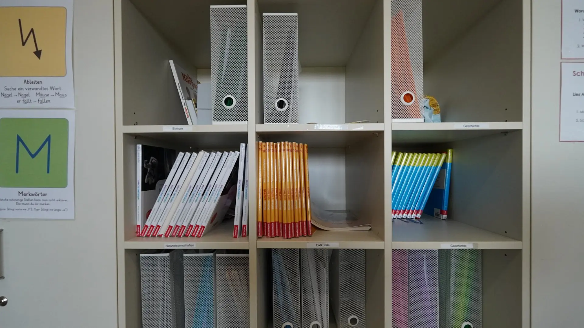 Gut sortiertes Materialregal mit Lehrbüchern für verschiedene Unterrichtsfächer und flankierenden Lernhilfen zur Sprachförderung an der Wand.