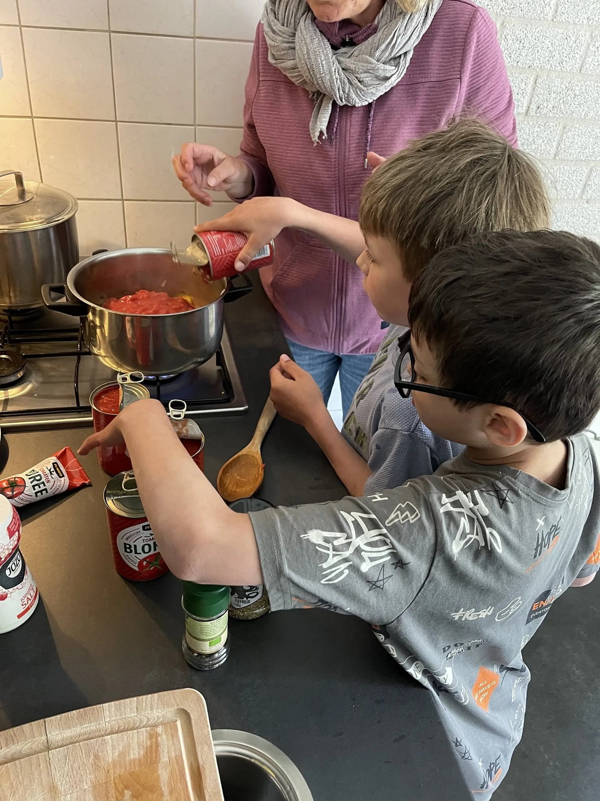 An einem Herd kochen zwei Kinder und eine Erwachsene Tomatensoße