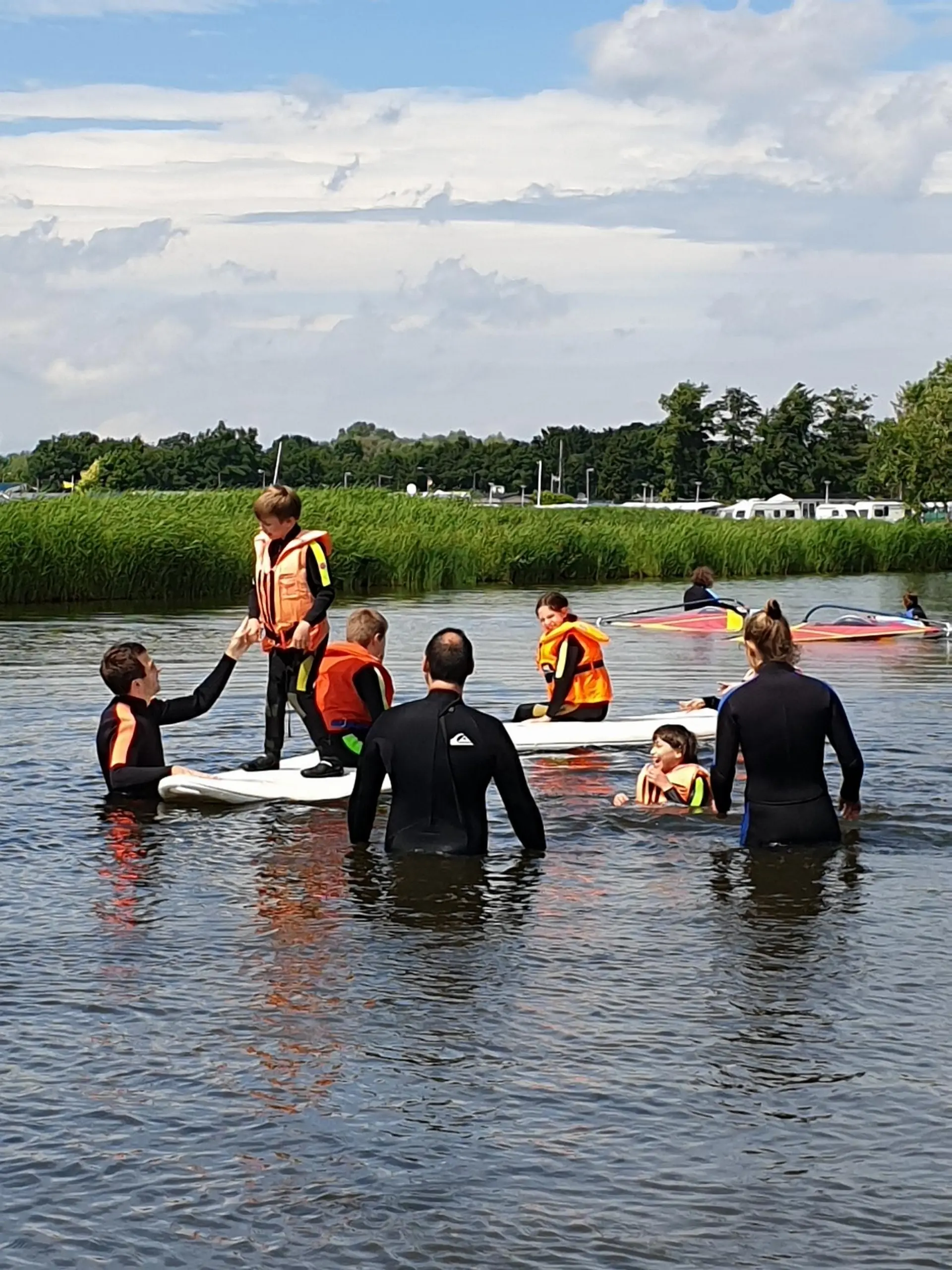 Kinder und Erwachsene in Neoprenanzügen üben im seichten Wasser mit Wind-Surf-Brettern
