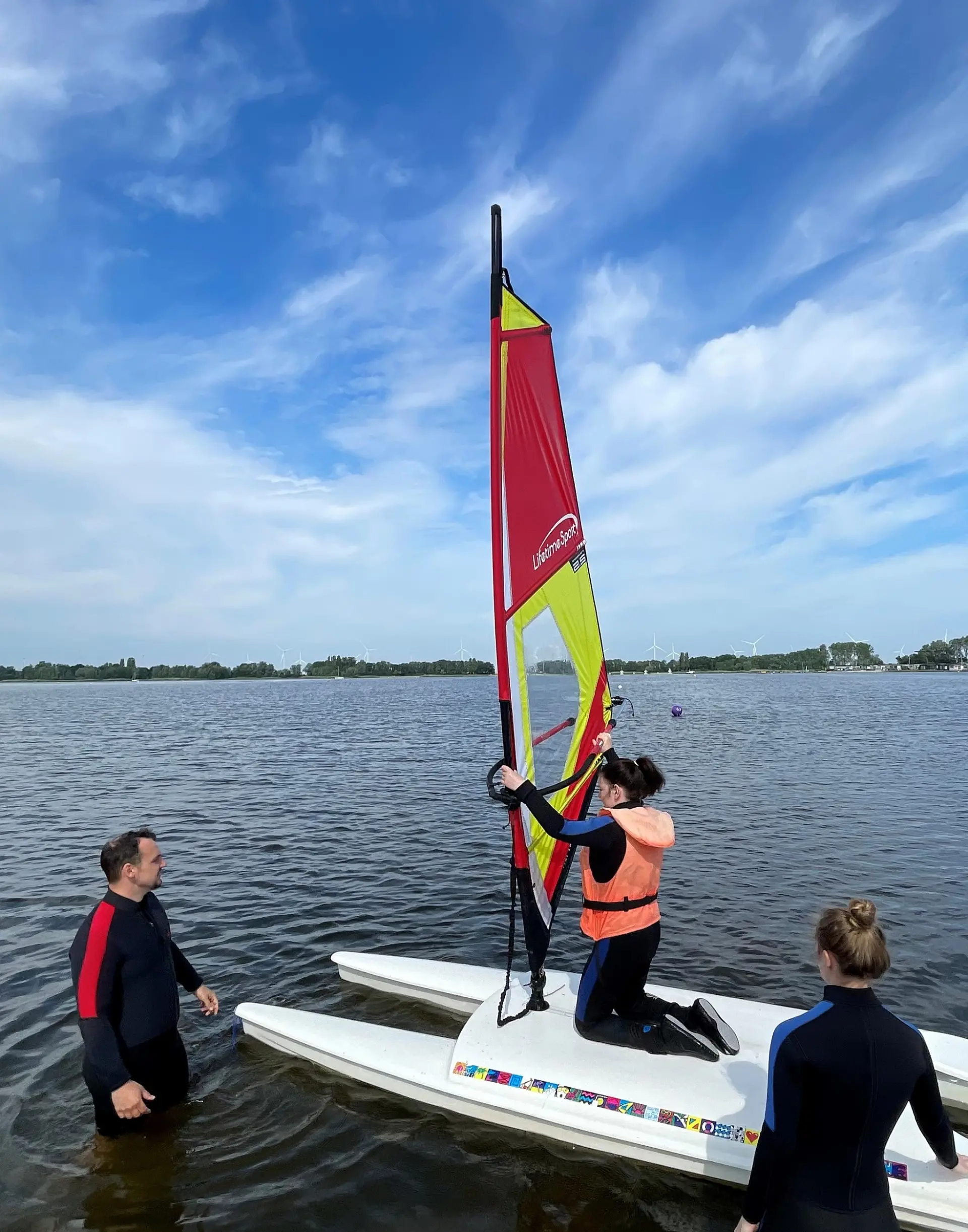 Zwei Erwachsene in Neoprenanzügen beobachten im seichten Wasser ein Kind, das auf einem Katamaran-artigem Surfbrett mit Segel kniet