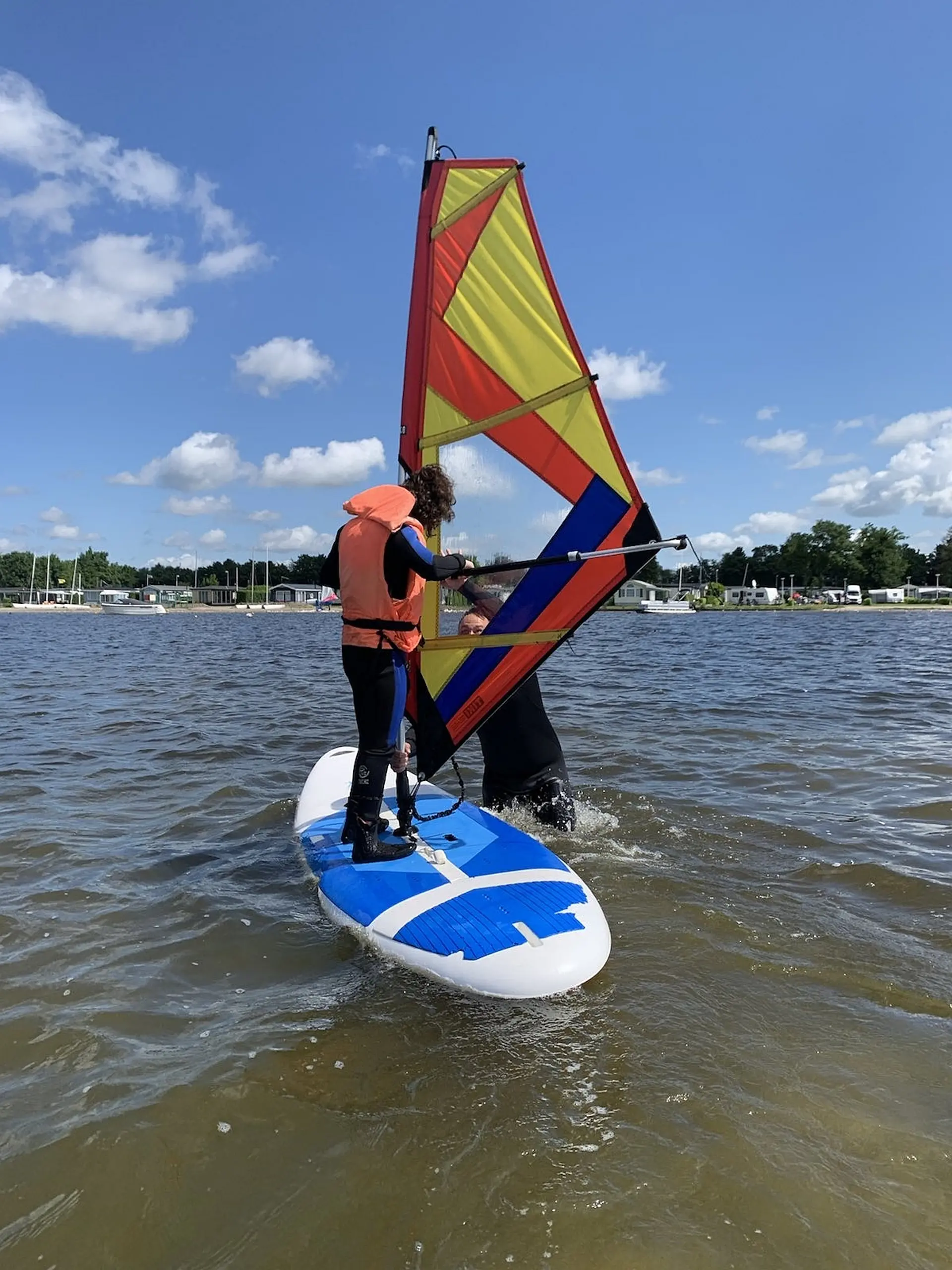 Ein Kind im Neoprenanzug steht auf einem Windsurfbrett, ein Erwachsener steht im Wasser und hält das Segel 