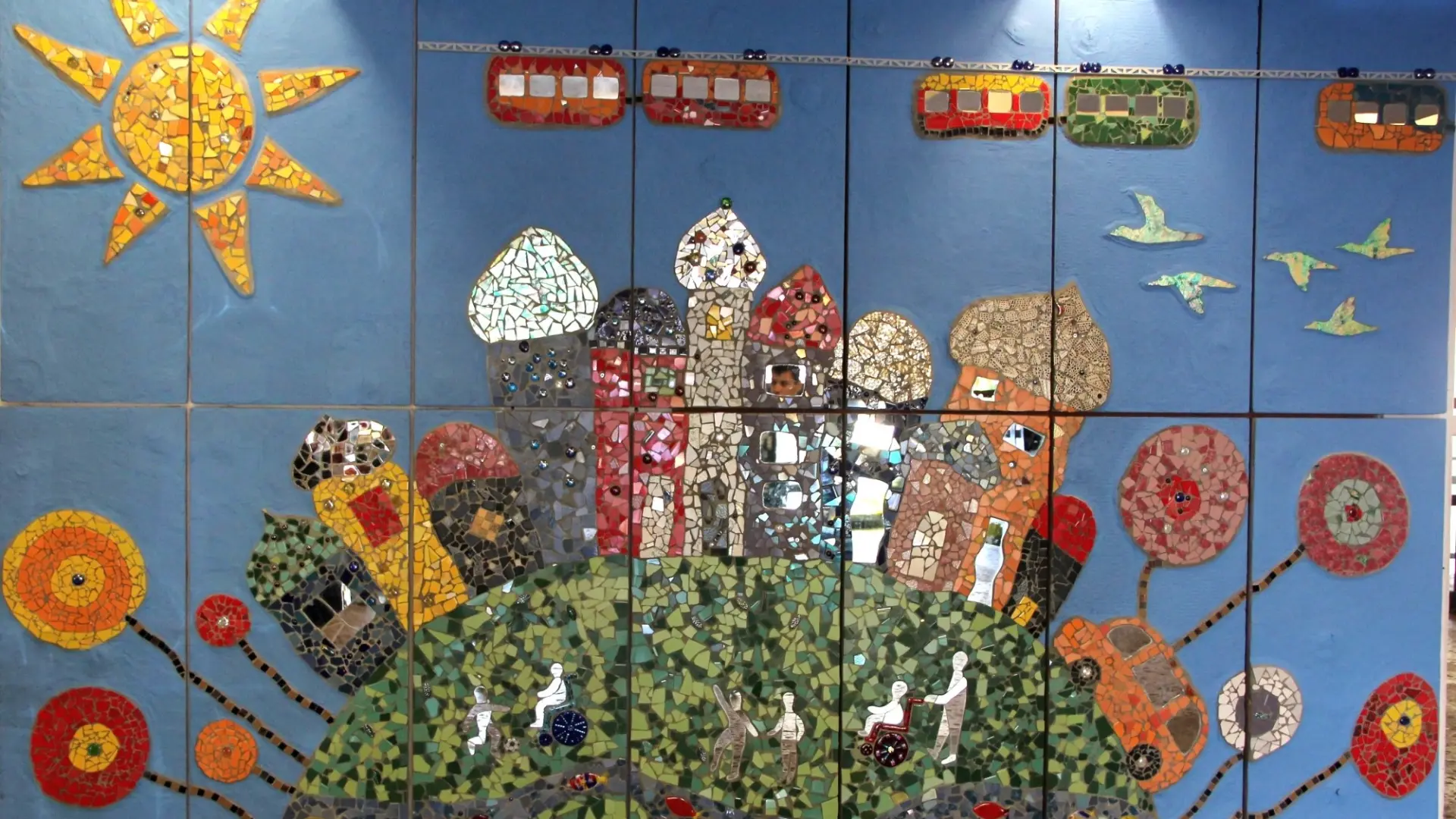 Ein Mosaik aus bunten Fliesen zeigt Sonne, Blumen, Häuser: davor Menschen mit und ohne Rollstuhl. Über allem fährt die Wuppertaler Schwebebahn.