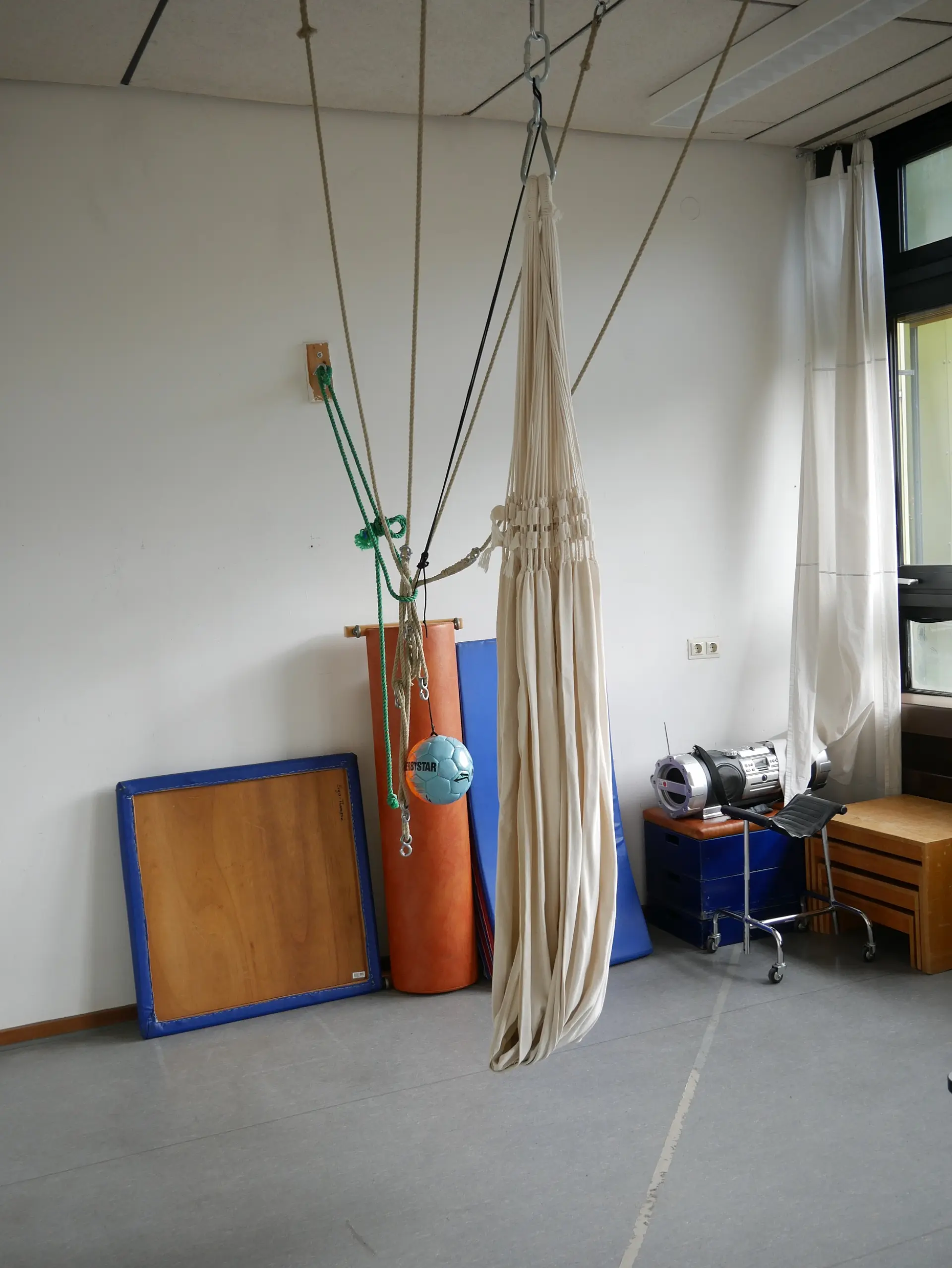 Eine weiße Tuchhängematte hängt von der Decke eines Therapieraums. Im Hintergrund stehen blaue Matten, eine orangefarbene Rolle und ein hängender Ball.