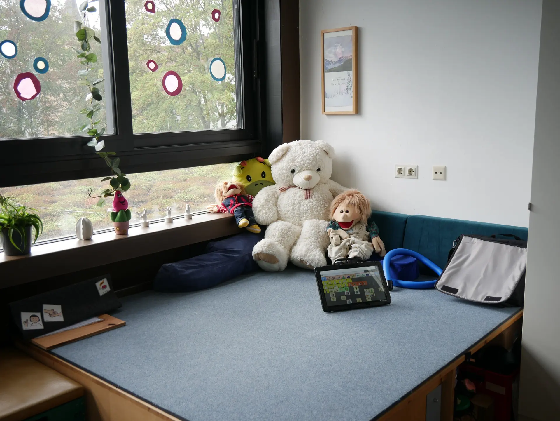 Gemütliche Sitzecke am Fenster mit vielen Kuscheltieren, darunter ein großer Eisbär. Auf der Polsterbank liegt ein Talker.