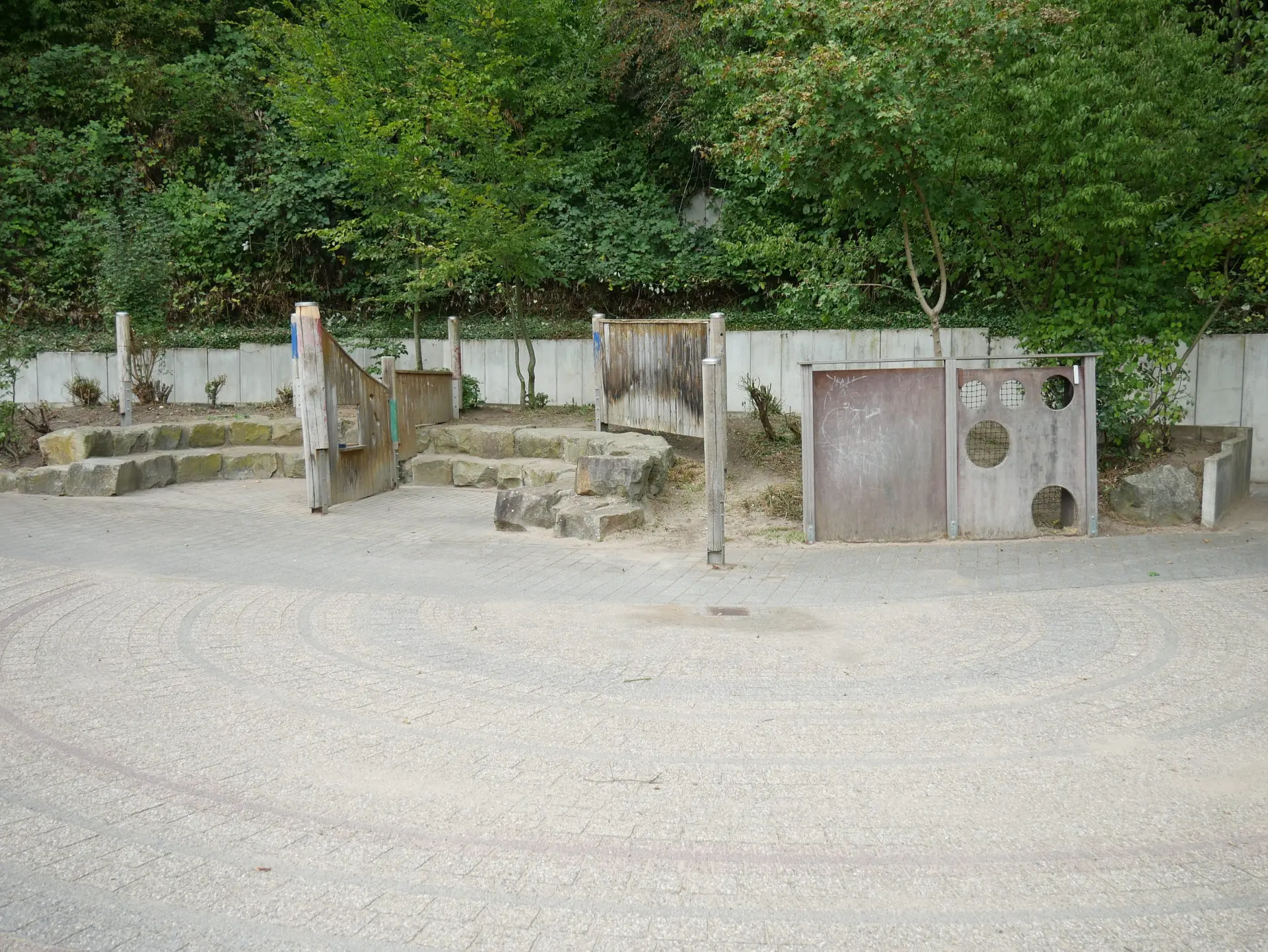 Ein gepflasterter Außenbereich mit Steinstufen und Holzelementen, umgeben von dichtem Grün. Der Boden zeigt kreisförmige Muster.