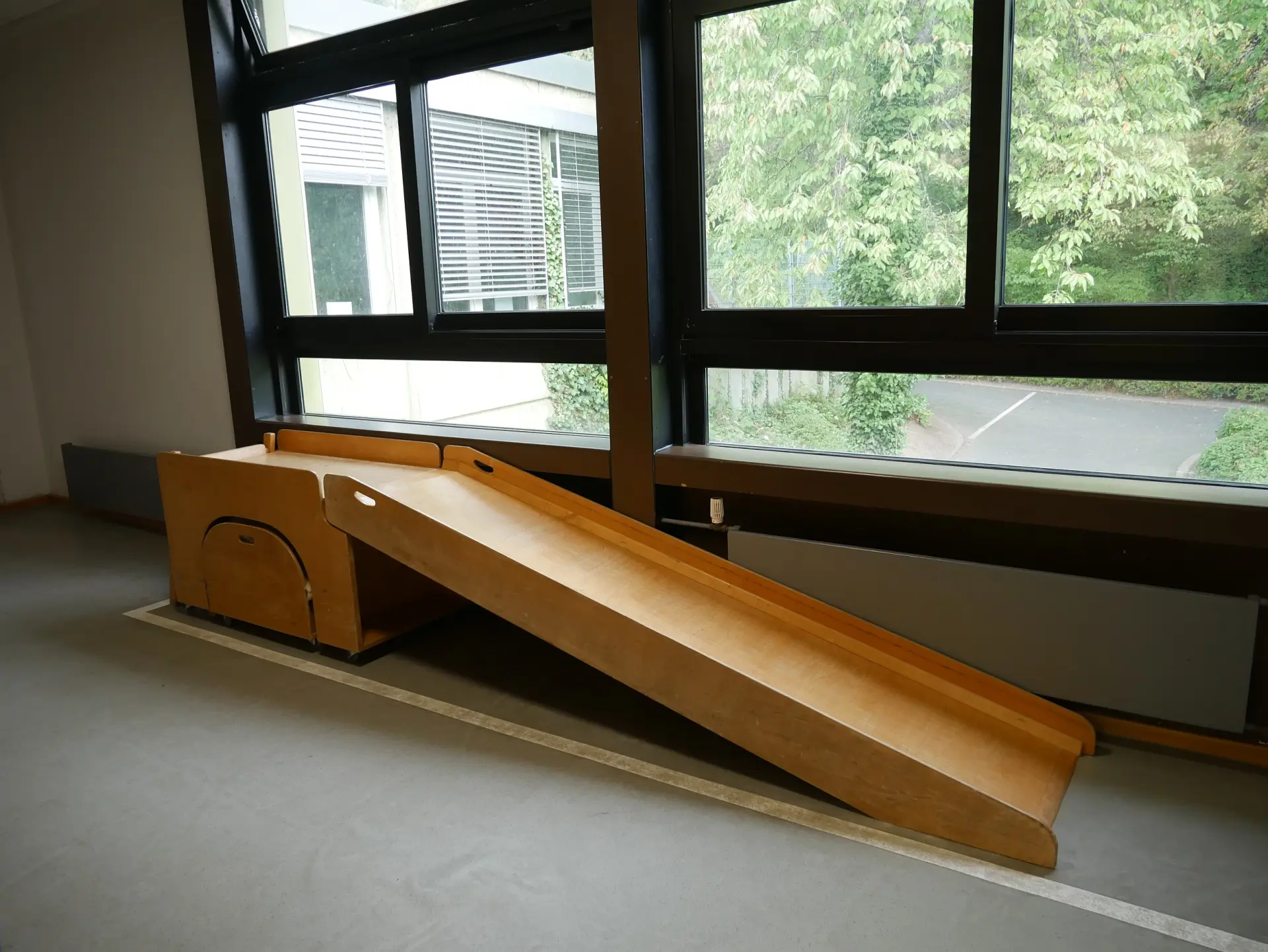 Eine kleine Kinderrutsche aus hellem Holz steht im Innenraum vor einem großen Fenster und führt auf den grauen Linoleumboden.