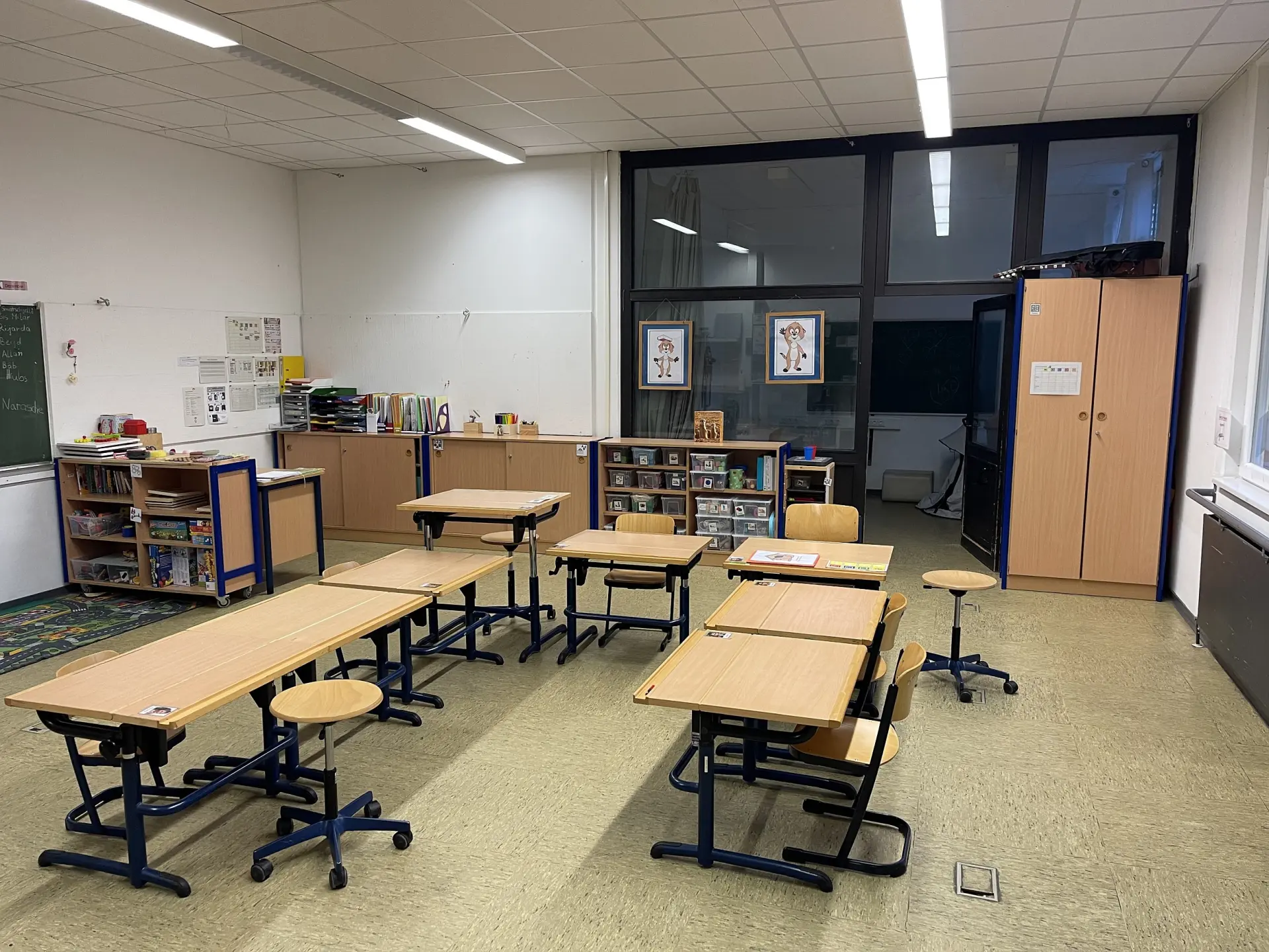 Ein helles Klassenzimmer mit Holzmöbeln. Schreibtische und Stühle sind in kleinen Gruppen arrangiert. Im Hintergrund stehen Regale und Schränke.