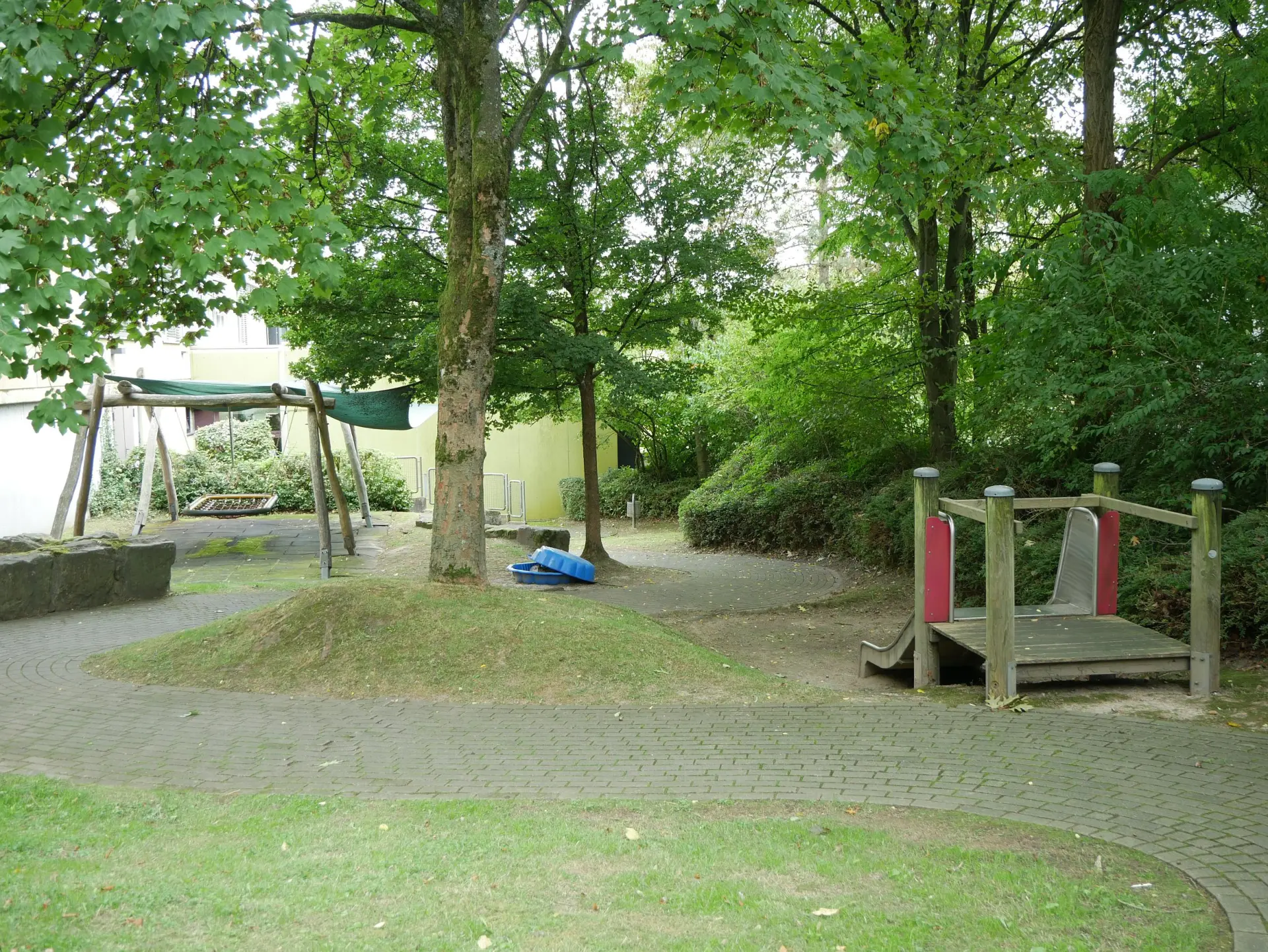 Ein Spielplatz im Freien mit einem gepflasterten Weg, einer Nestschaukel unter einem grünen Sonnensegel und einem kleinen Holz-Klettergerüst mit Rutsche.